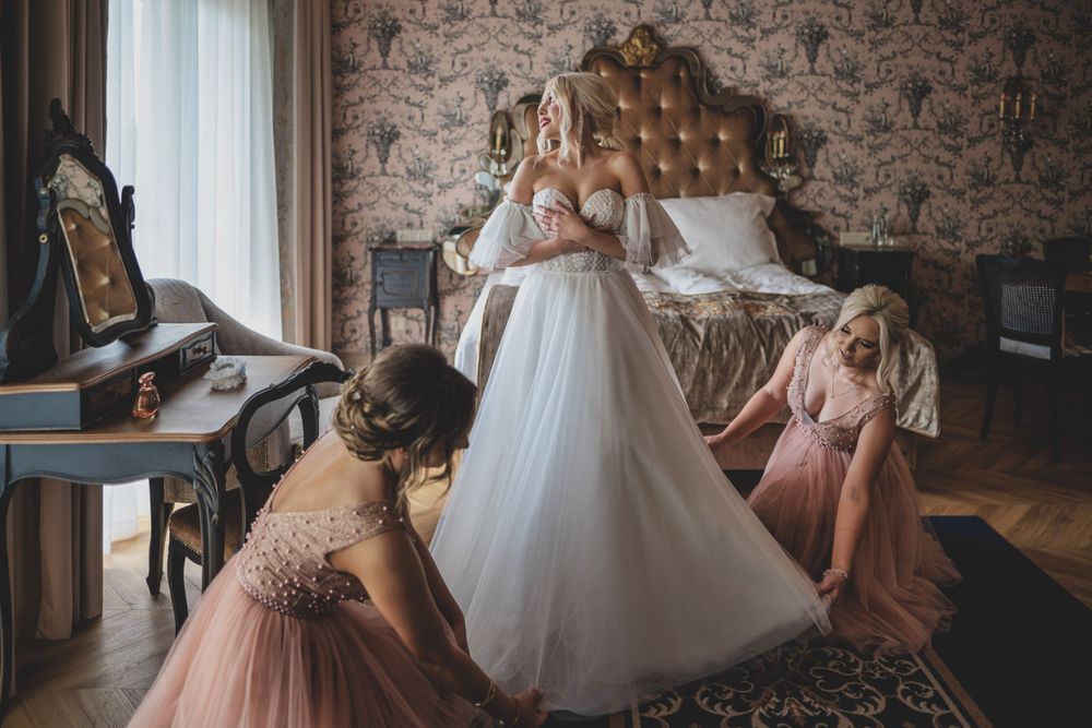Druhny w różowych sukienkach pomagają poprawić zwiewną białą suknię ślubną w eleganckiej sypialni w stylu vintage. Ślub w Piszu