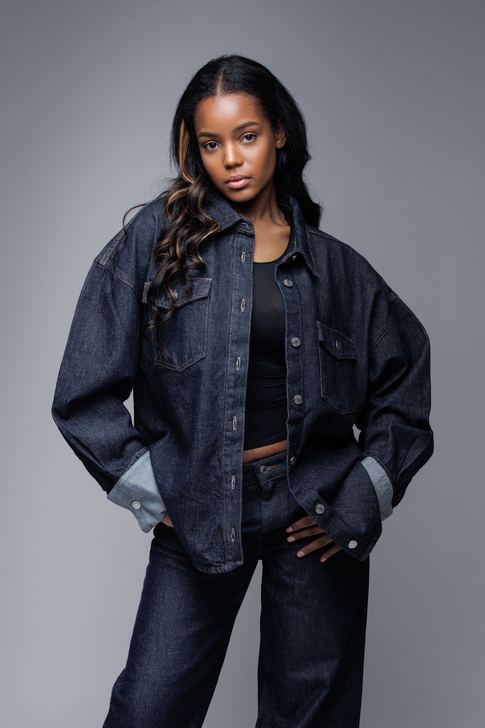 Model wearing oversized denim jacket and jeans in studio portrait against gray background.