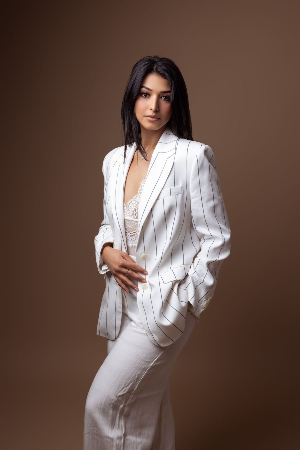 A person wearing an elegant white pinstriped pantsuit and lace top poses against a brown studio backdrop.