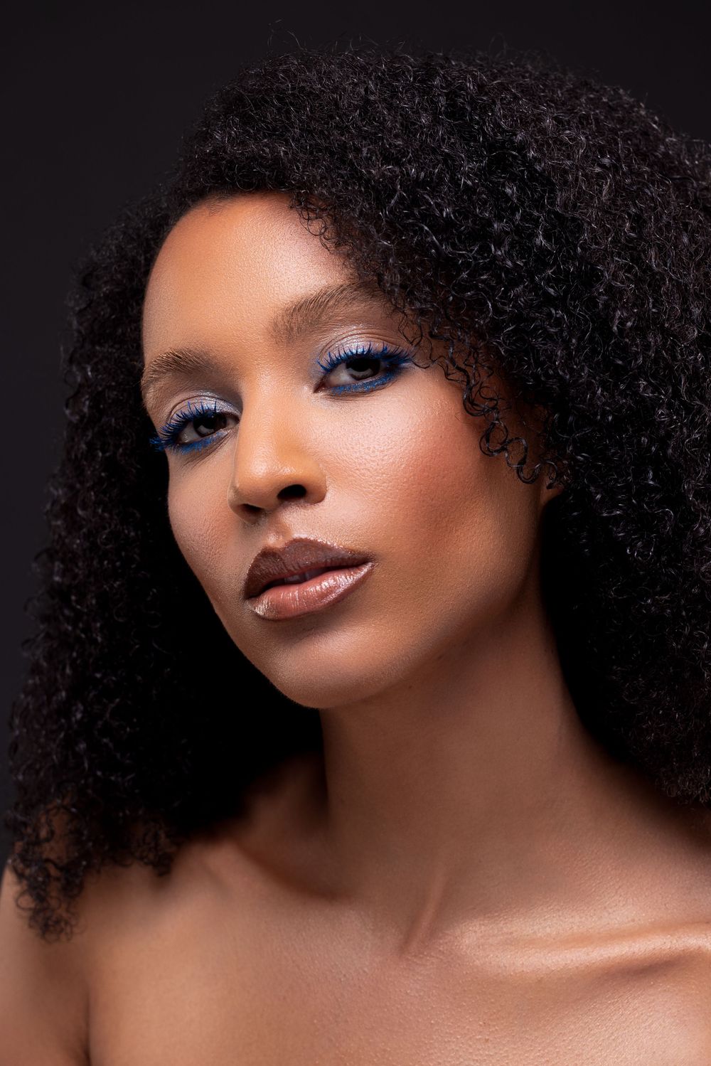 Close-up beauty portrait with blue eyeshadow, curly natural hair, and brown lipstick against dark background.