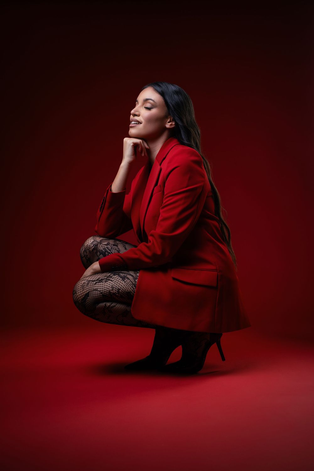 Fashion portrait against red background featuring model in bright red blazer and dark pants crouching in heels.