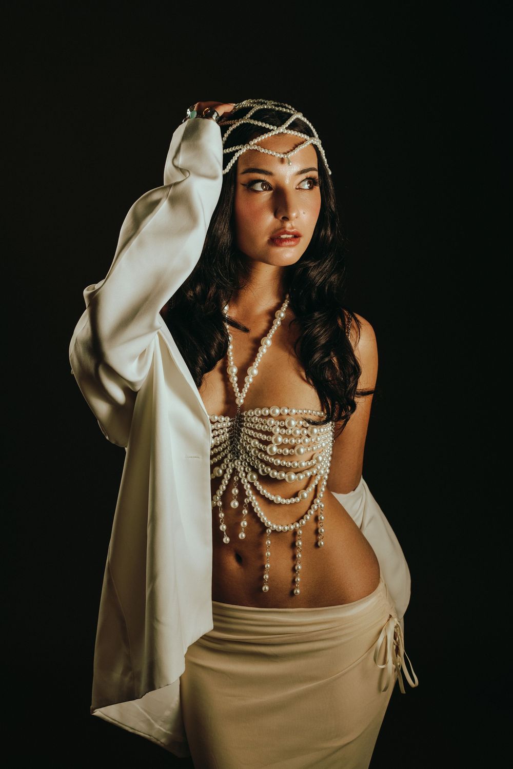 Woman in pearl bead top, head chain, white satin skirt and jacket poses against dark background.