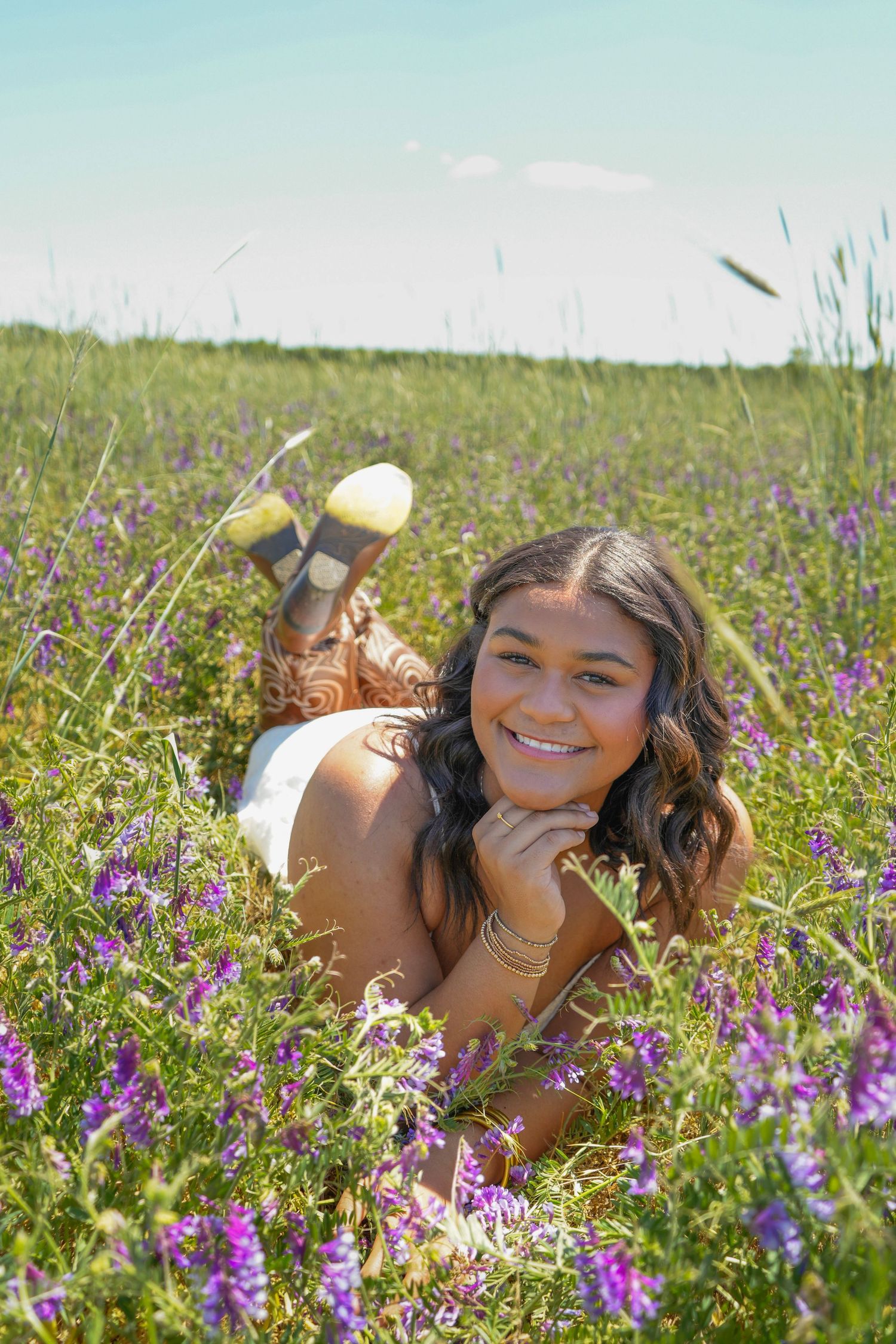 Smiling young woman lying in a field of purple wildflowers on a sunny day, wearing a white dress and yellow shoes.