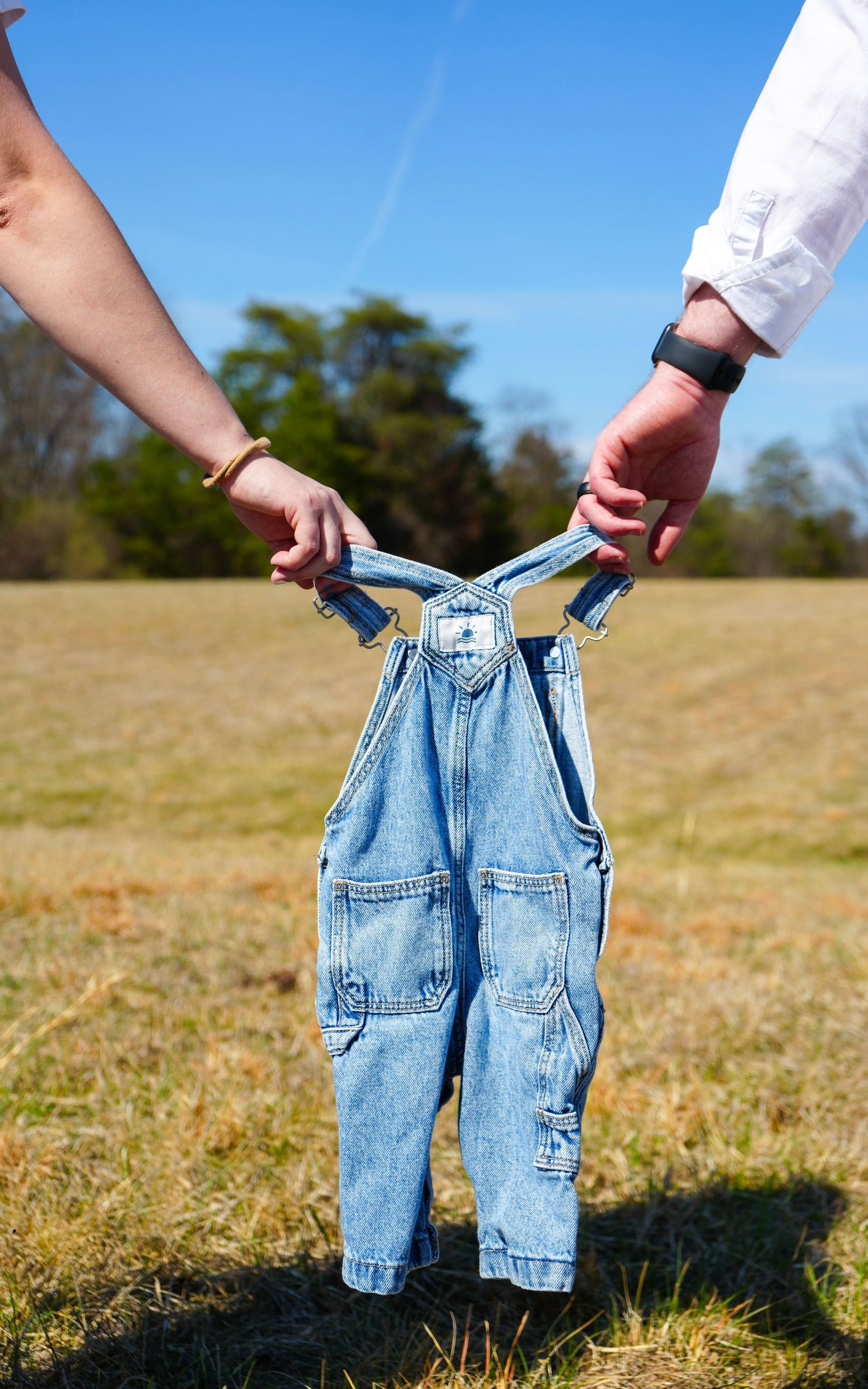 Two people holding tiny denim overalls outdoors in a field, a sweet pregnancy announcement photo.