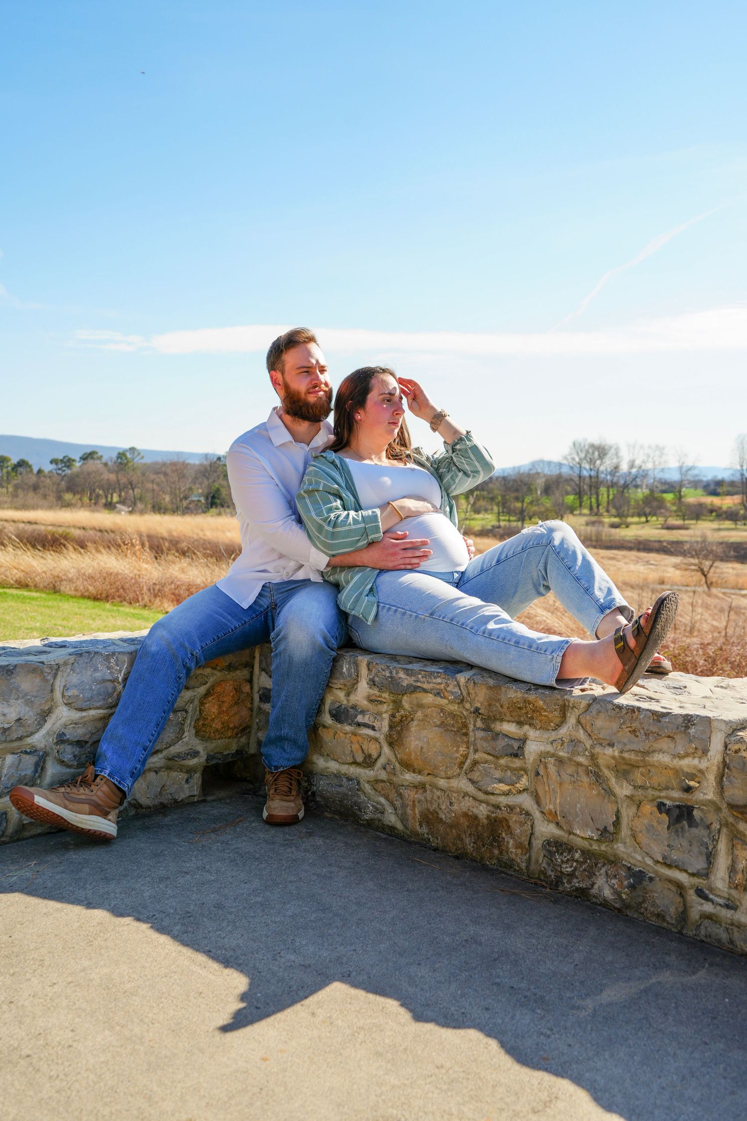 A couple sits on a stone wall outdoors, the woman reclining against the man, both in jeans, with a scenic landscape behind.