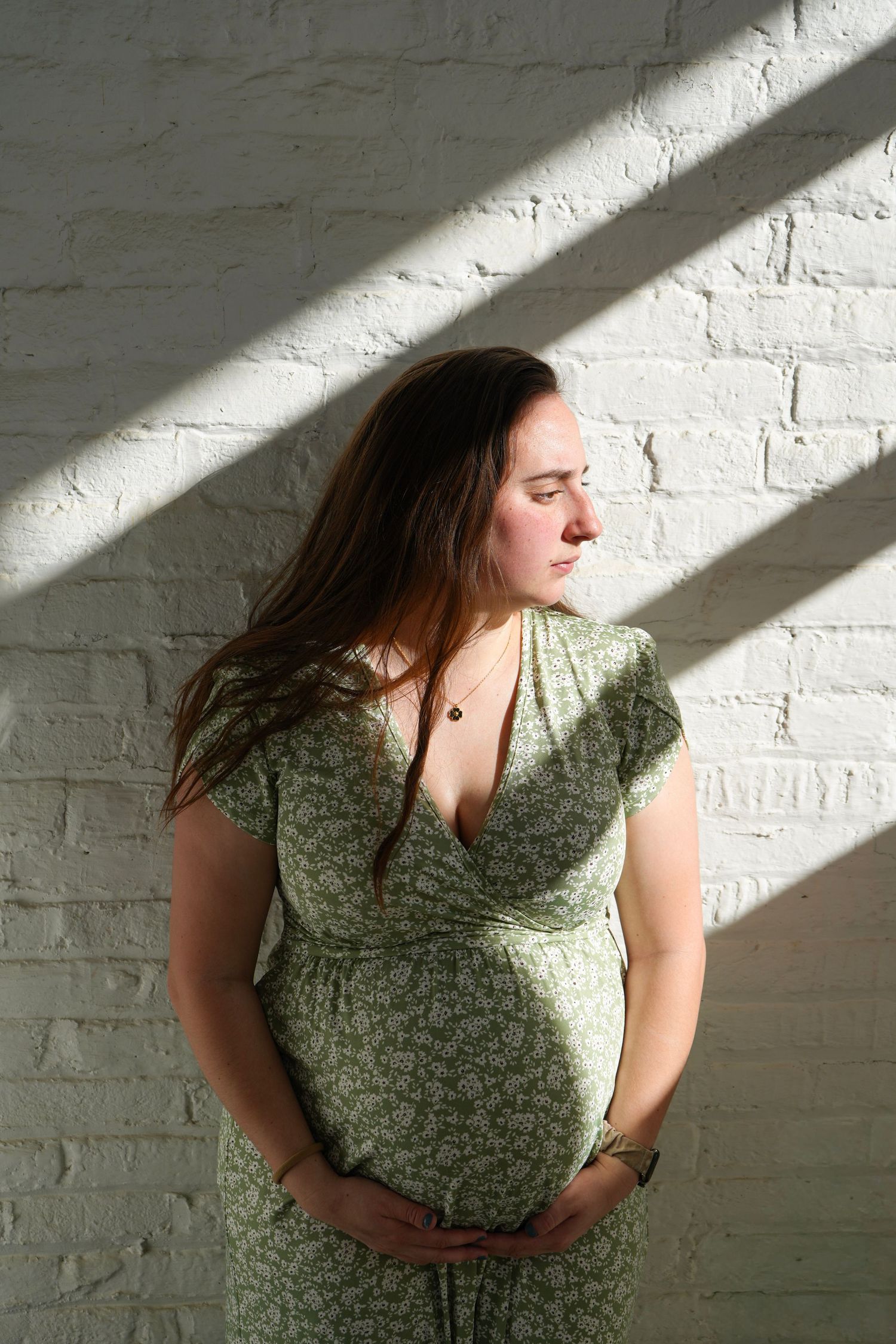 Pregnant woman in green floral dress cradling baby bump against white brick wall in dramatic sunlight.