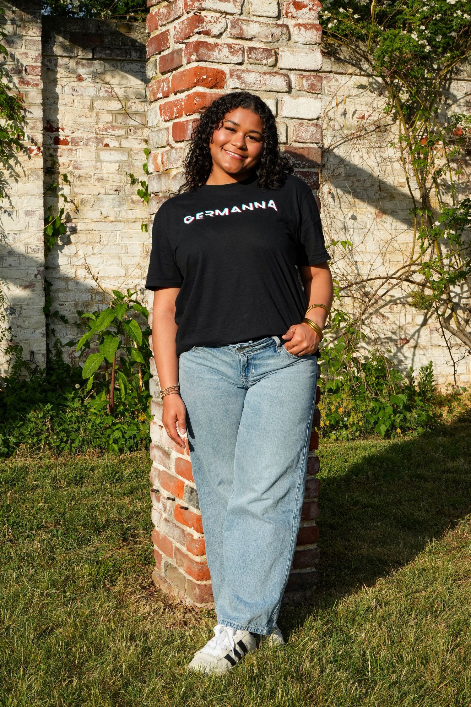 Woman wearing black graphic tee and light wash jeans standing outdoors against brick wall in natural sunlight.