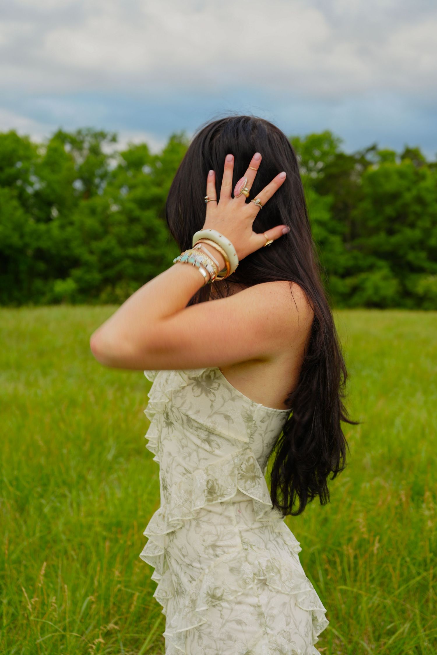 Woman in white ruffled dress with bangles raises hand to face in lush green field under cloudy sky.