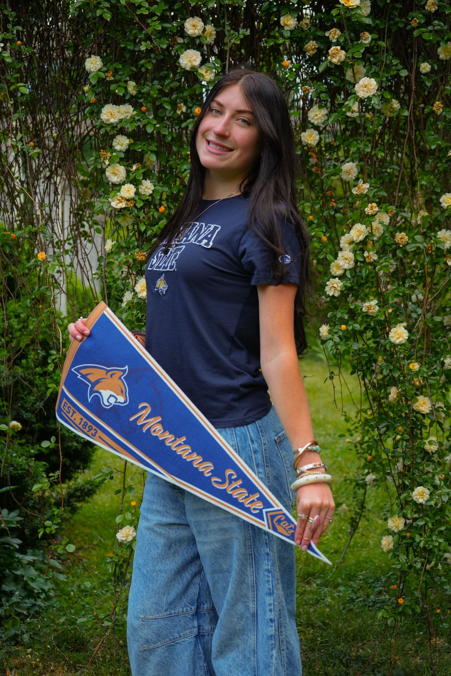 Smiling young woman holding a Montana State University pennant flag, wearing a navy MSU t-shirt outdoors.