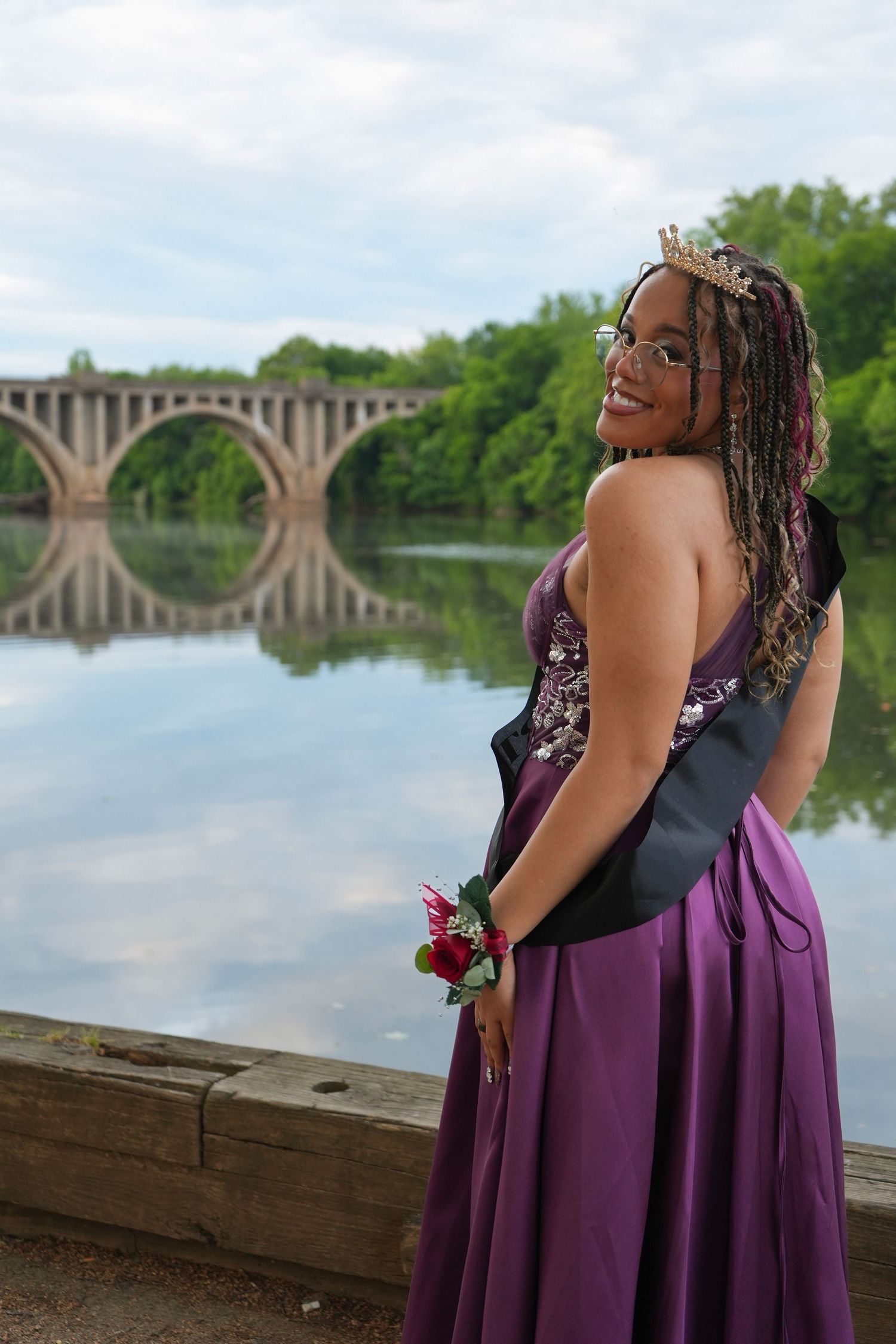 Young woman in purple prom dress with corsage and tiara poses by a river with bridge backdrop.