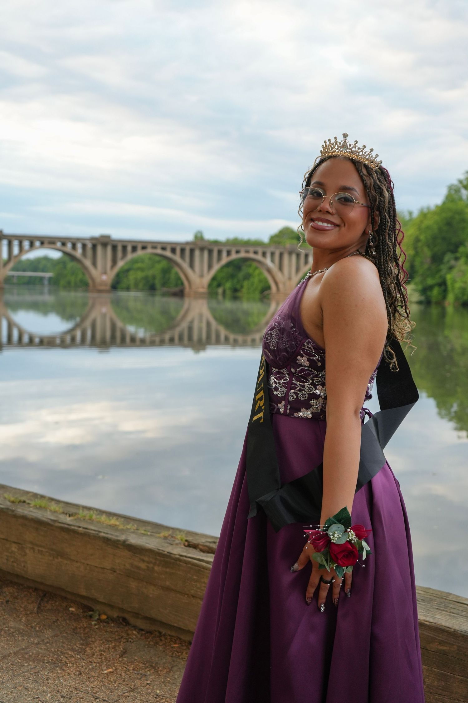 Young woman in purple prom dress and tiara poses by a river with an arched bridge backdrop.