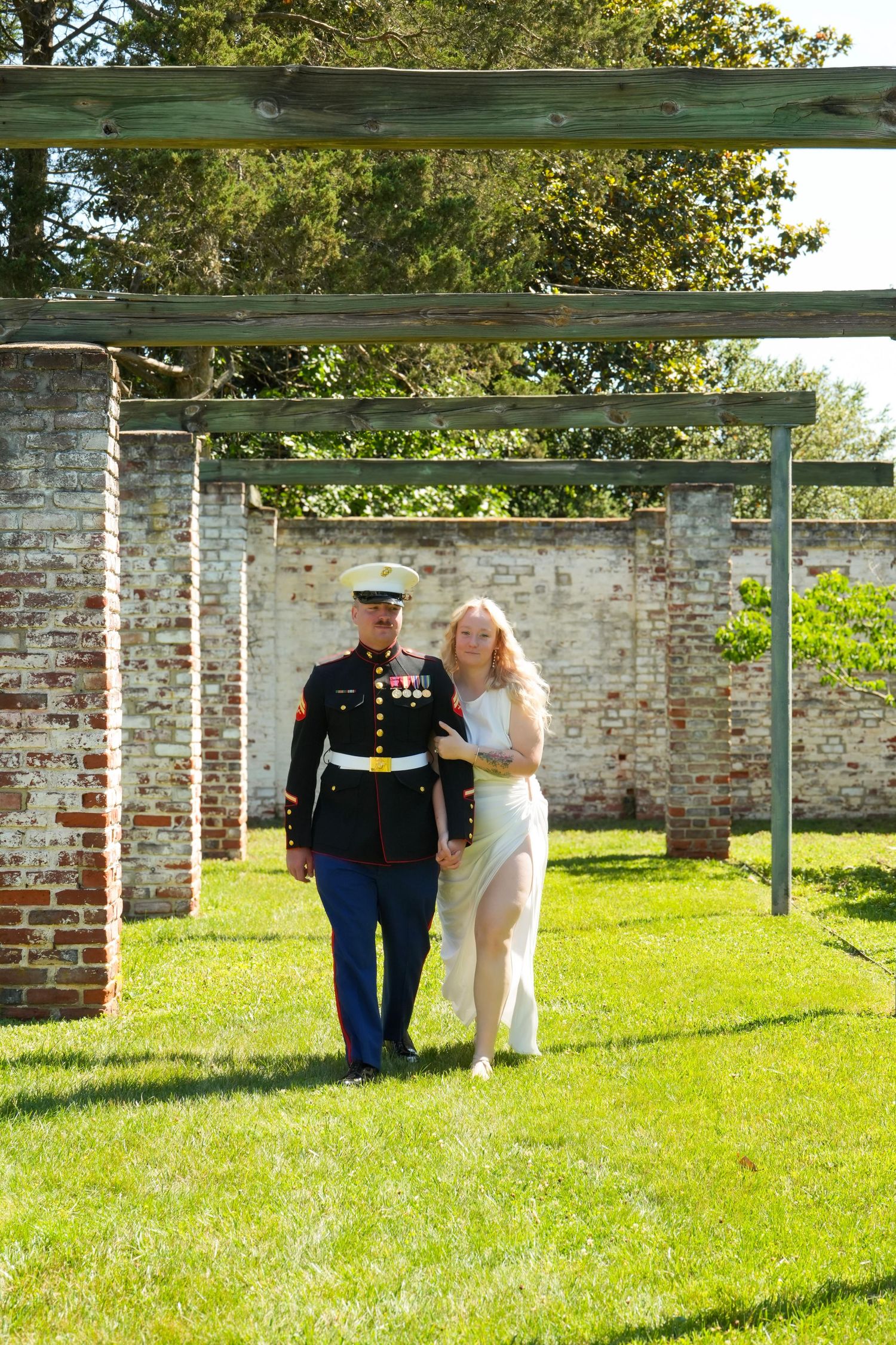 Marine in dress uniform walks with blonde woman in white dress on green lawn before brick wall and wooden pergola.