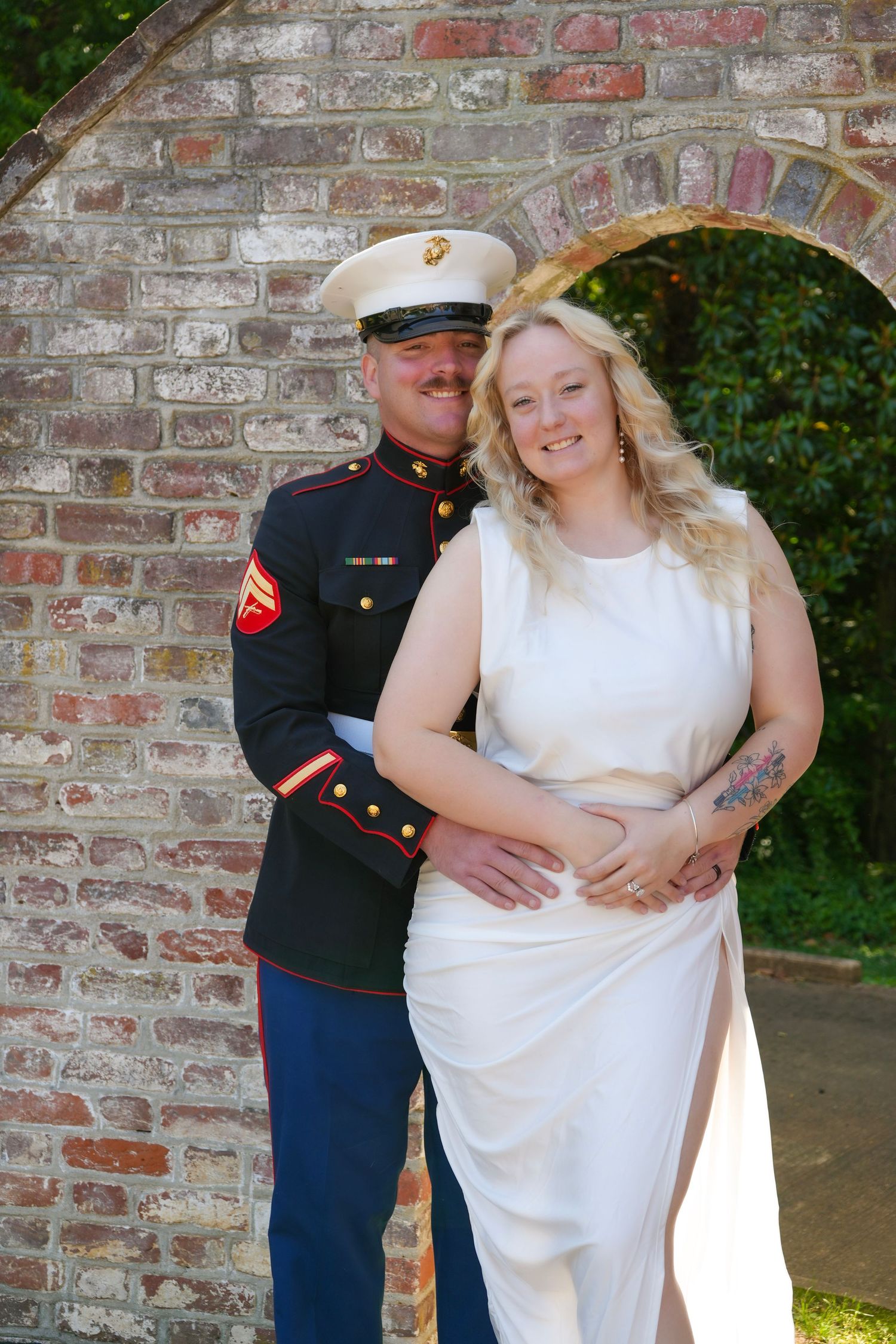 A US Marine in dress uniform poses with a smiling blonde bride in white dress before a brick archway.