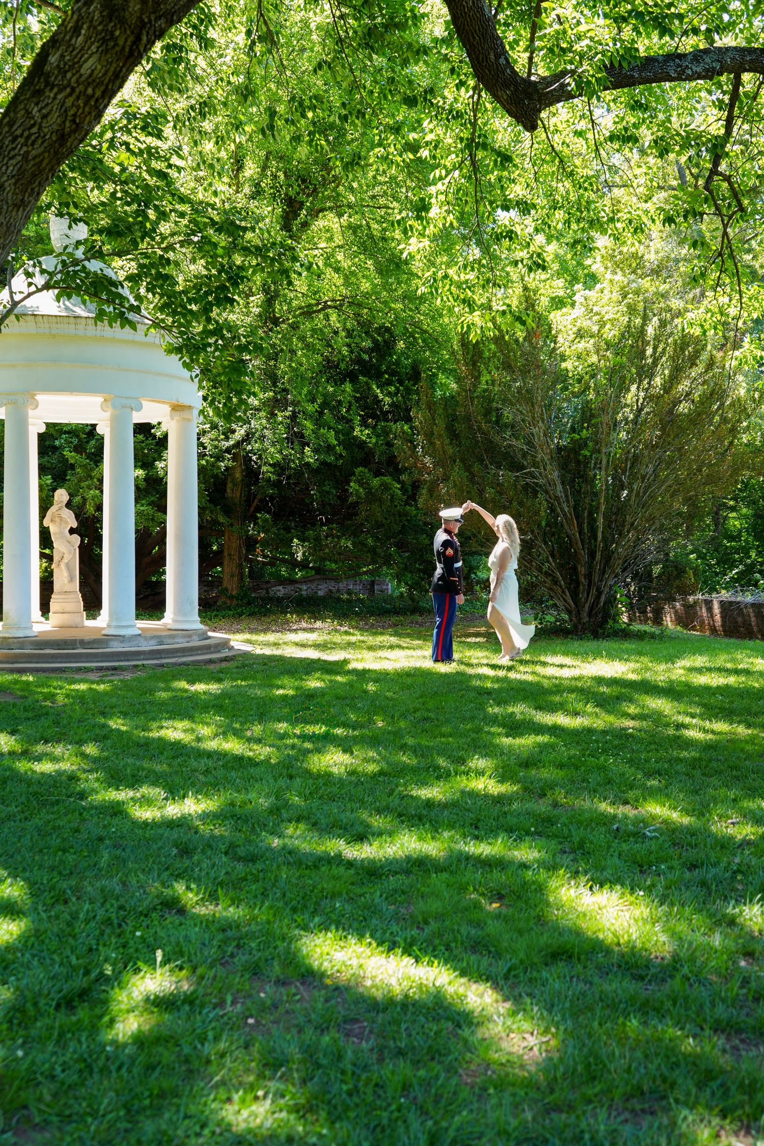 Bride and groom dancing on lush green lawn near white gazebo surrounded by tall trees on wedding day.