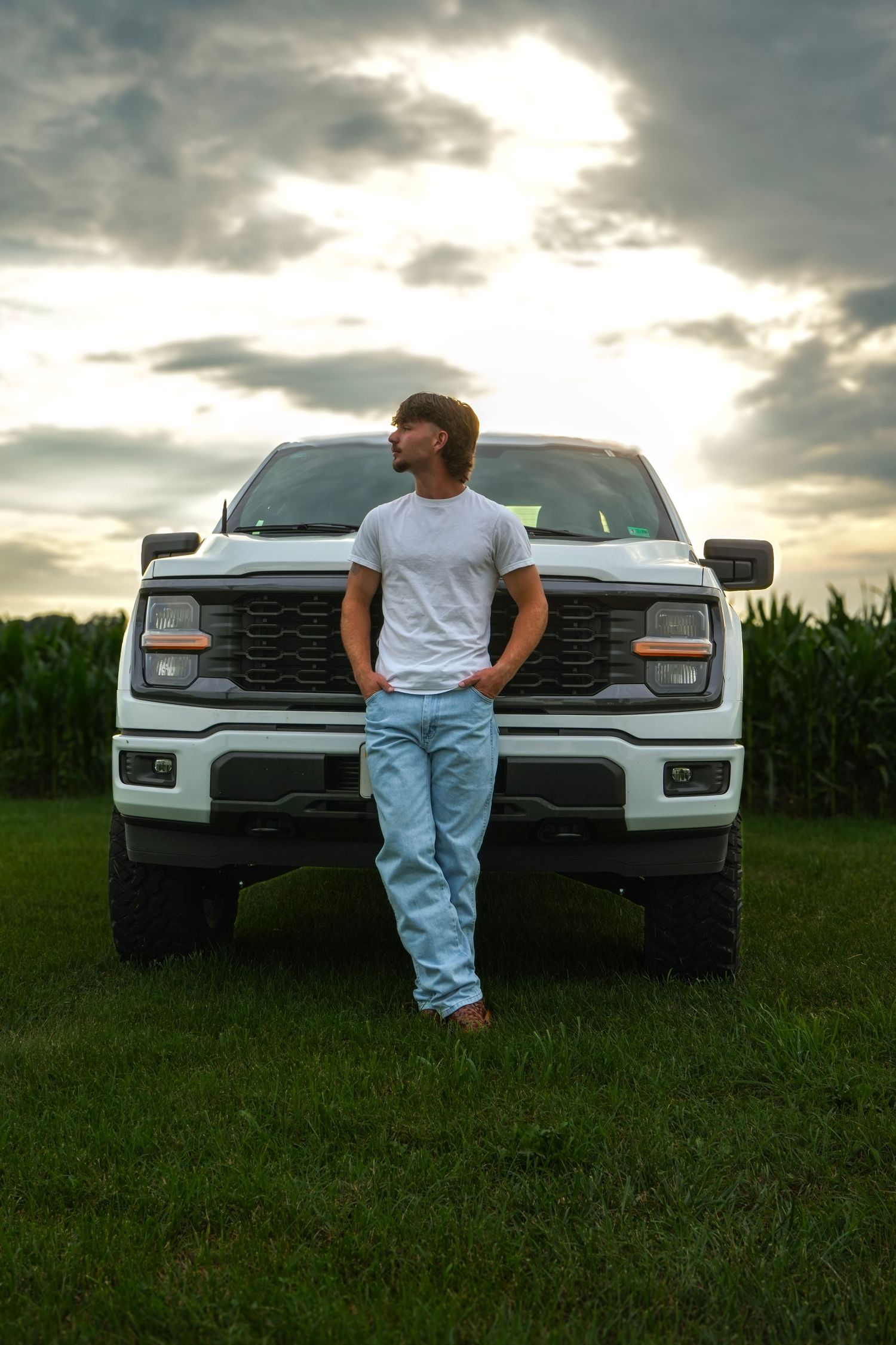Young man in white tee and jeans leaning against lifted white pickup truck in rural field at sunset.