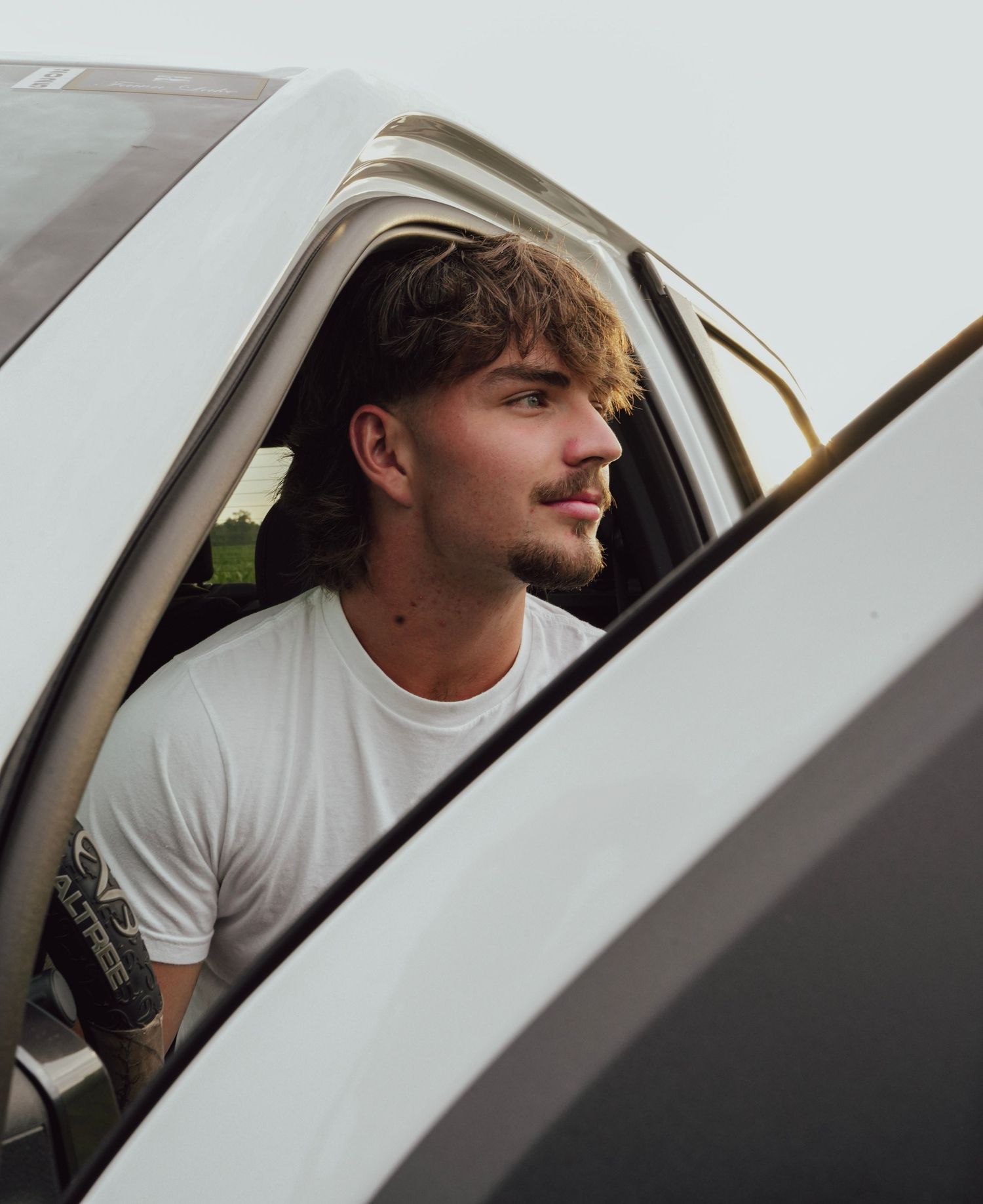 Young man with beard in white t-shirt sitting in car doorway looking away, casual outdoor portrait.
