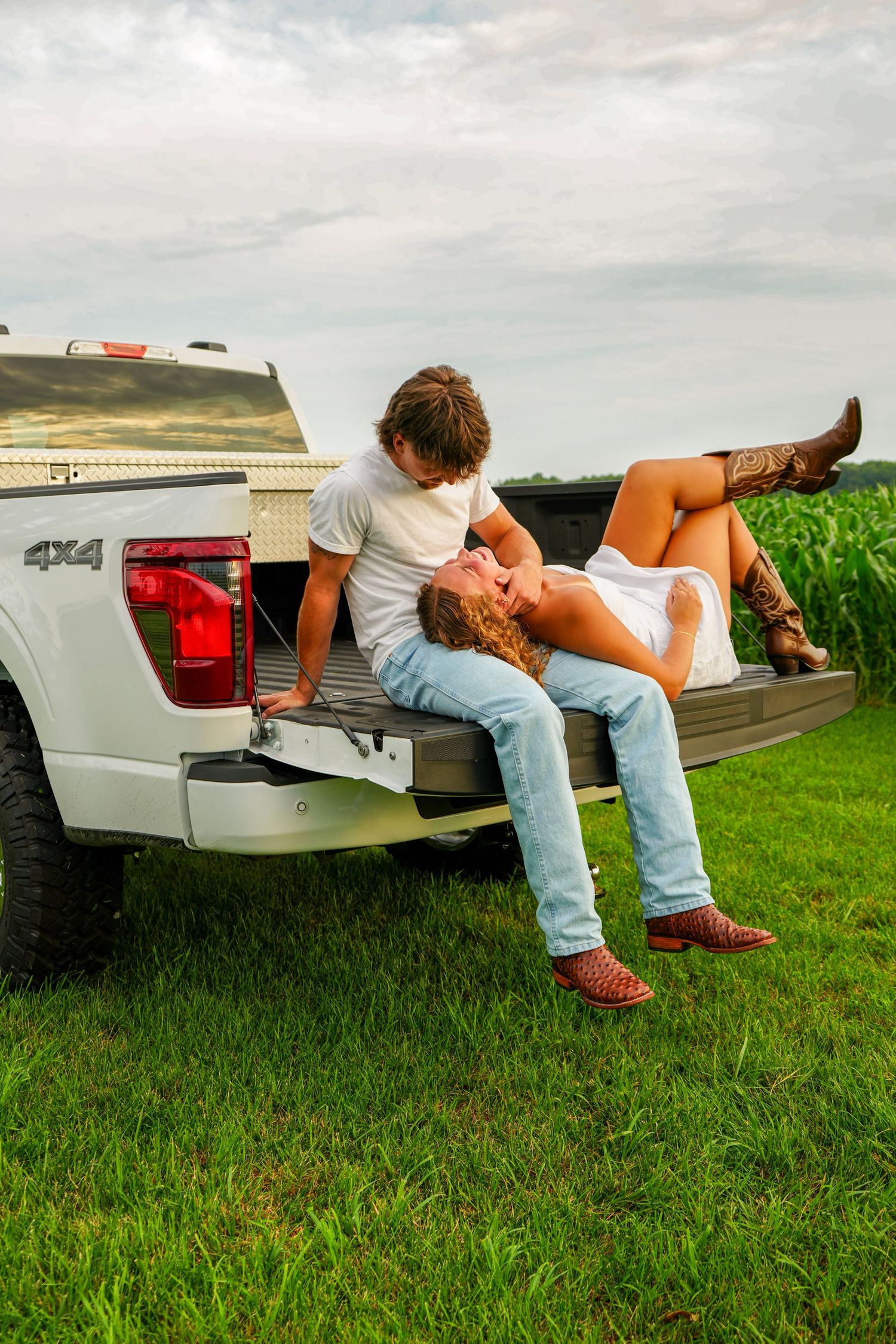 Couple sharing a romantic kiss on the tailgate of a white 4x4 pickup truck near a cornfield.