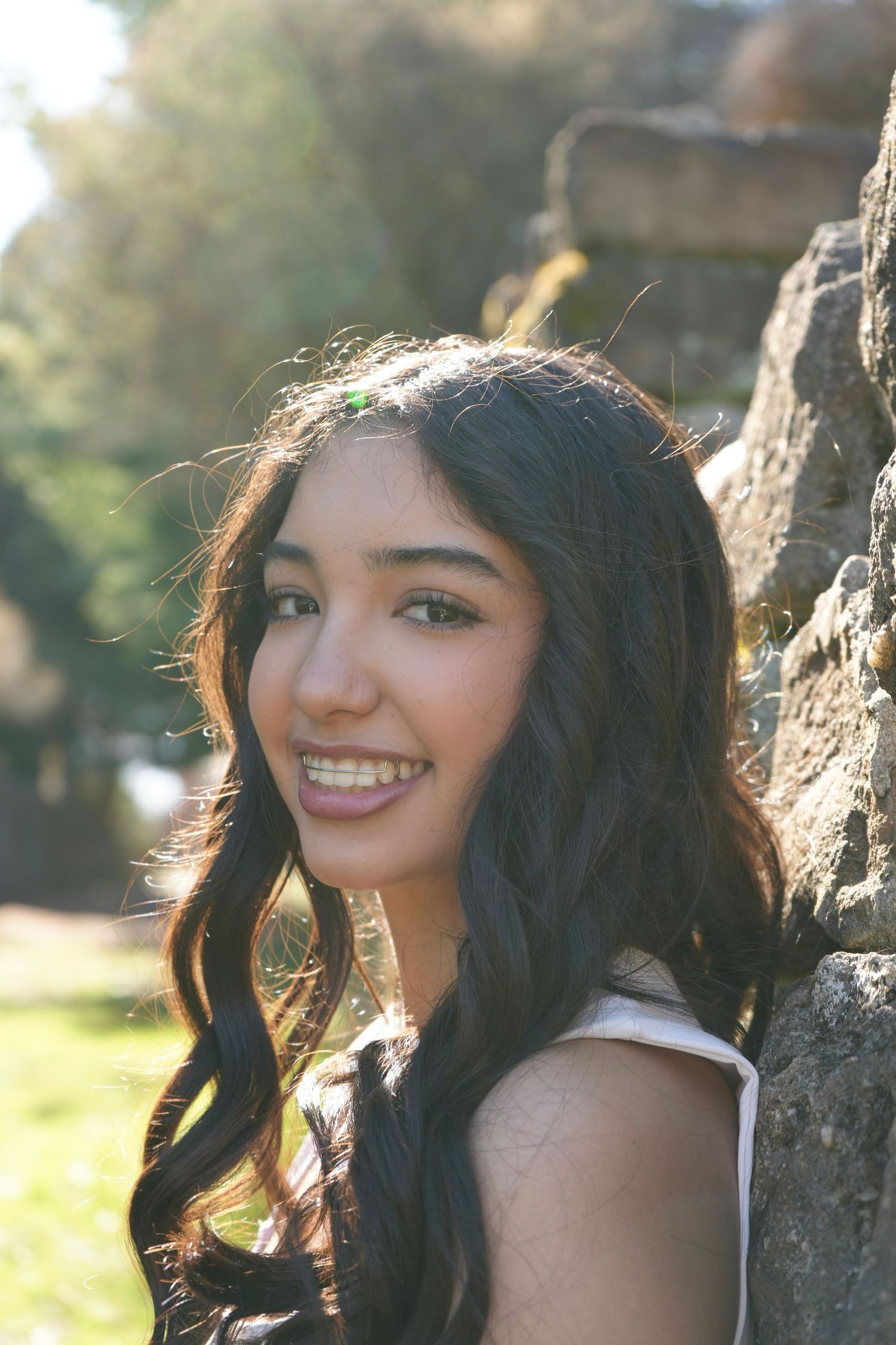 Smiling young woman with long dark wavy hair leaning against a stone wall outdoors in sunlight.