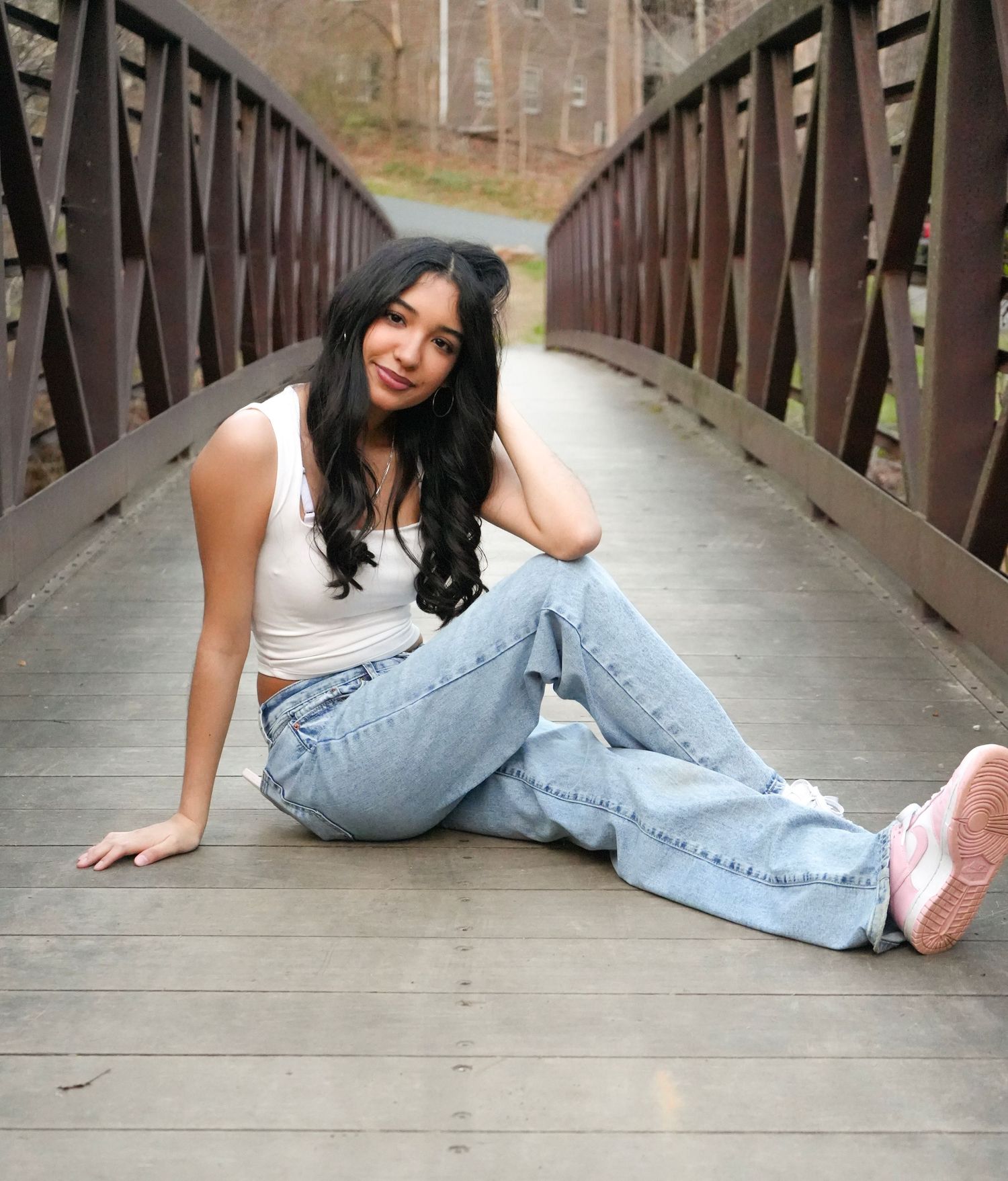 Young woman in white crop top and light wash flare jeans sitting on a wooden bridge, smiling with pink sneakers.