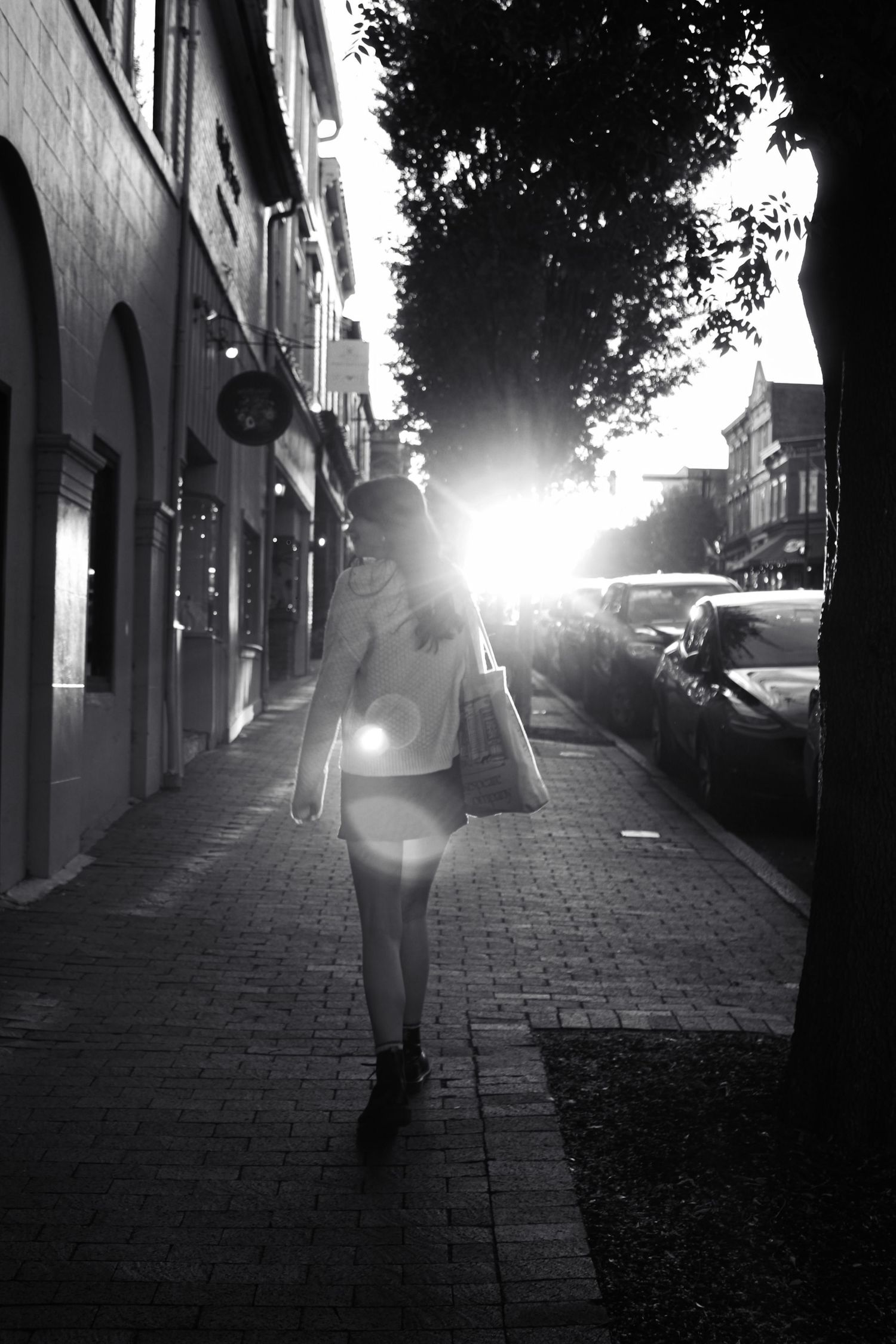 Black and white photo of a woman walking on a brick sidewalk toward bright sunlight, with shops and parked cars nearby.