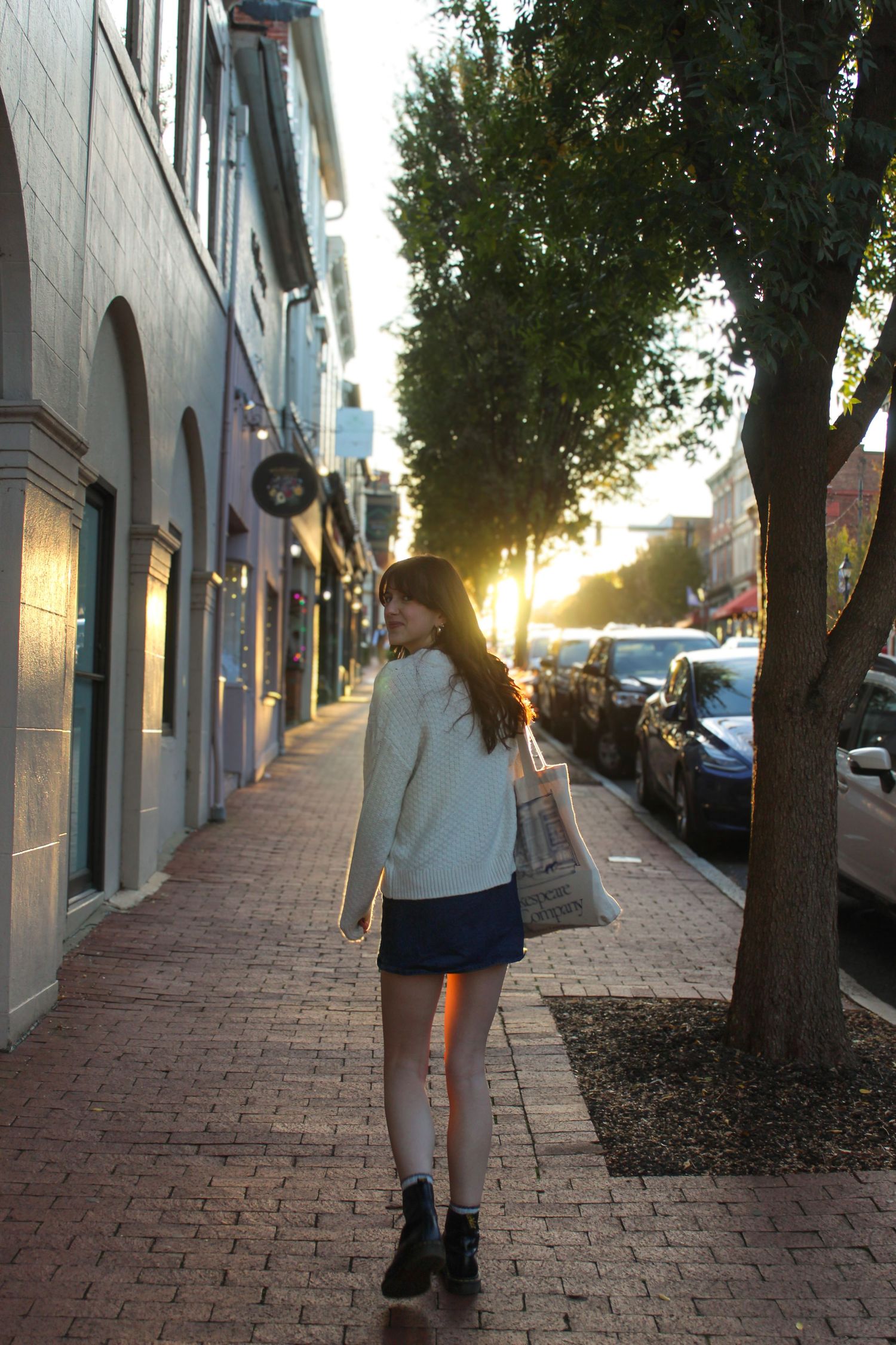 Young woman in white sweater and mini skirt walking on brick sidewalk at golden hour sunset.