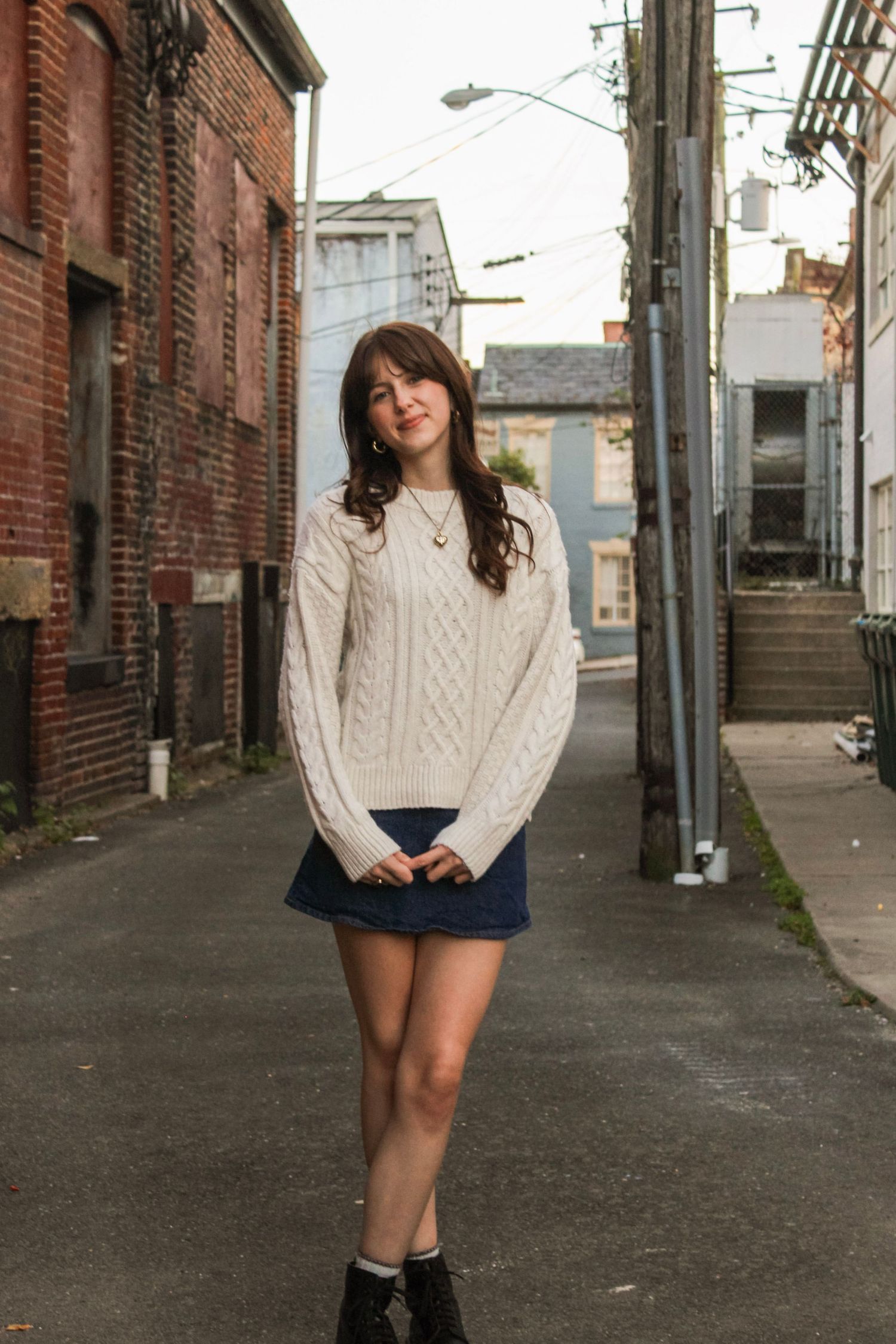 Young woman in white cable-knit sweater and denim mini skirt standing in urban alley between brick buildings.