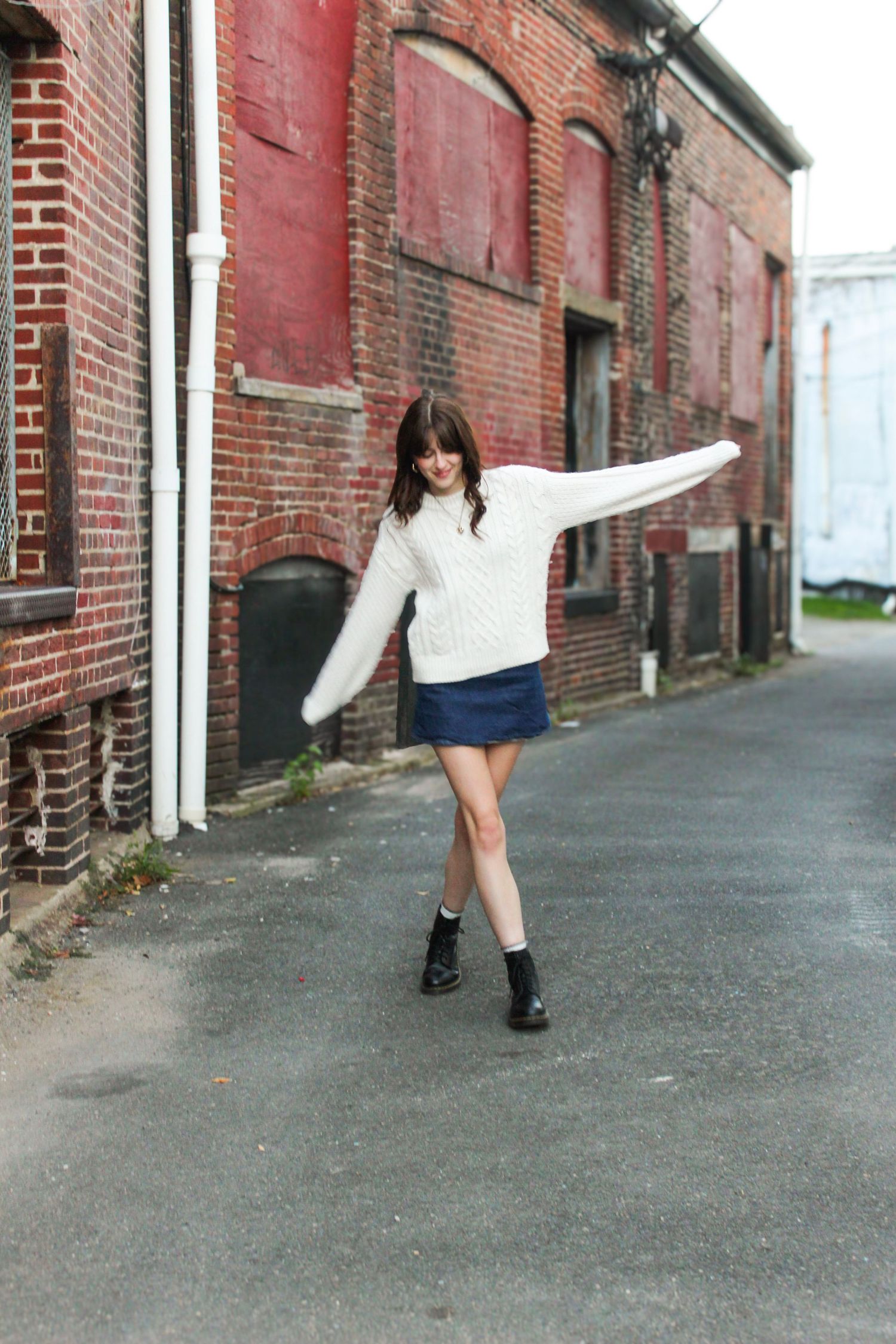 Young woman in white cable knit sweater and denim mini skirt poses in urban alley with red brick buildings.