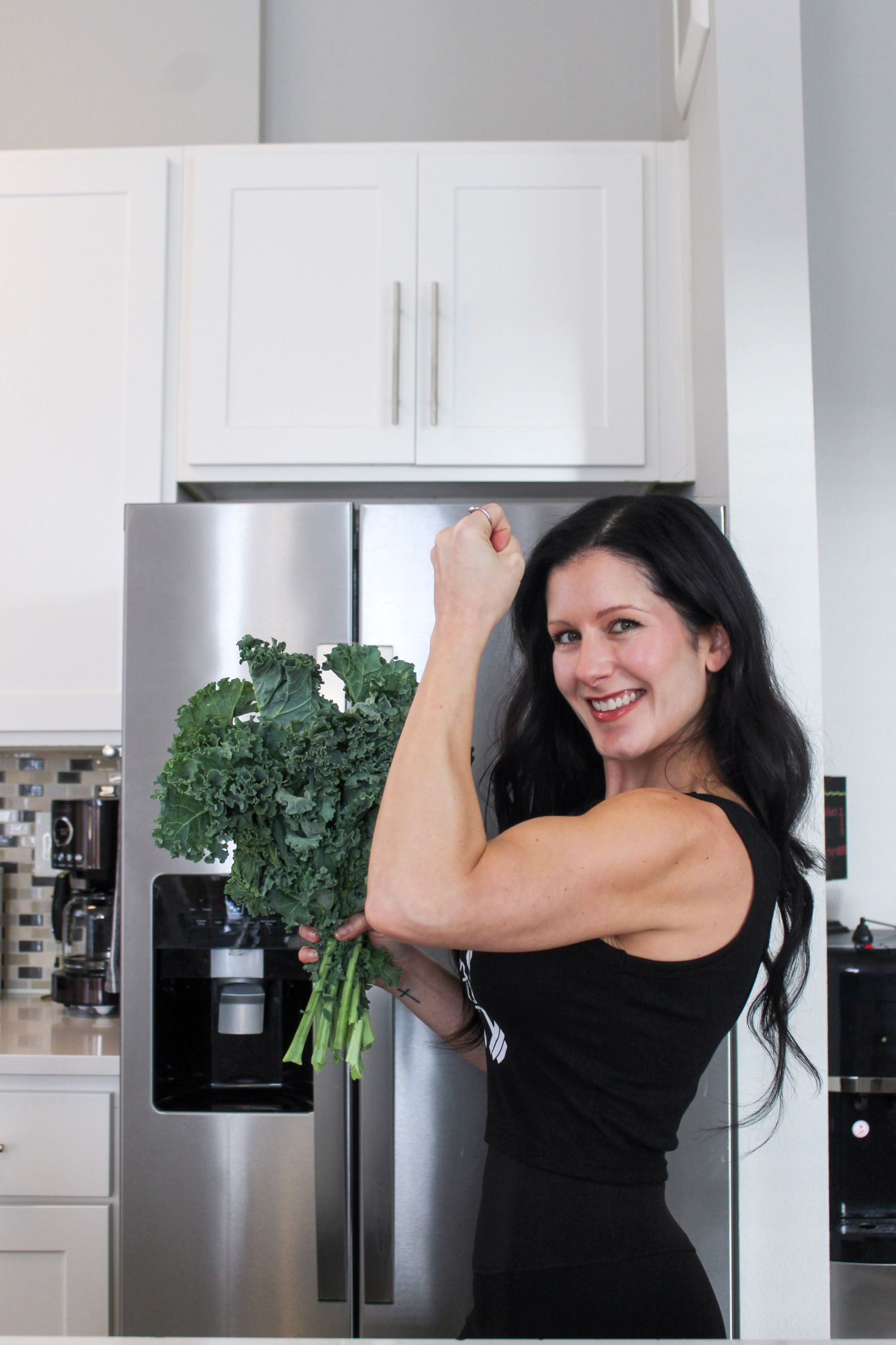 Fit woman flexing her bicep while holding fresh kale in a modern white kitchen with stainless steel refrigerator.