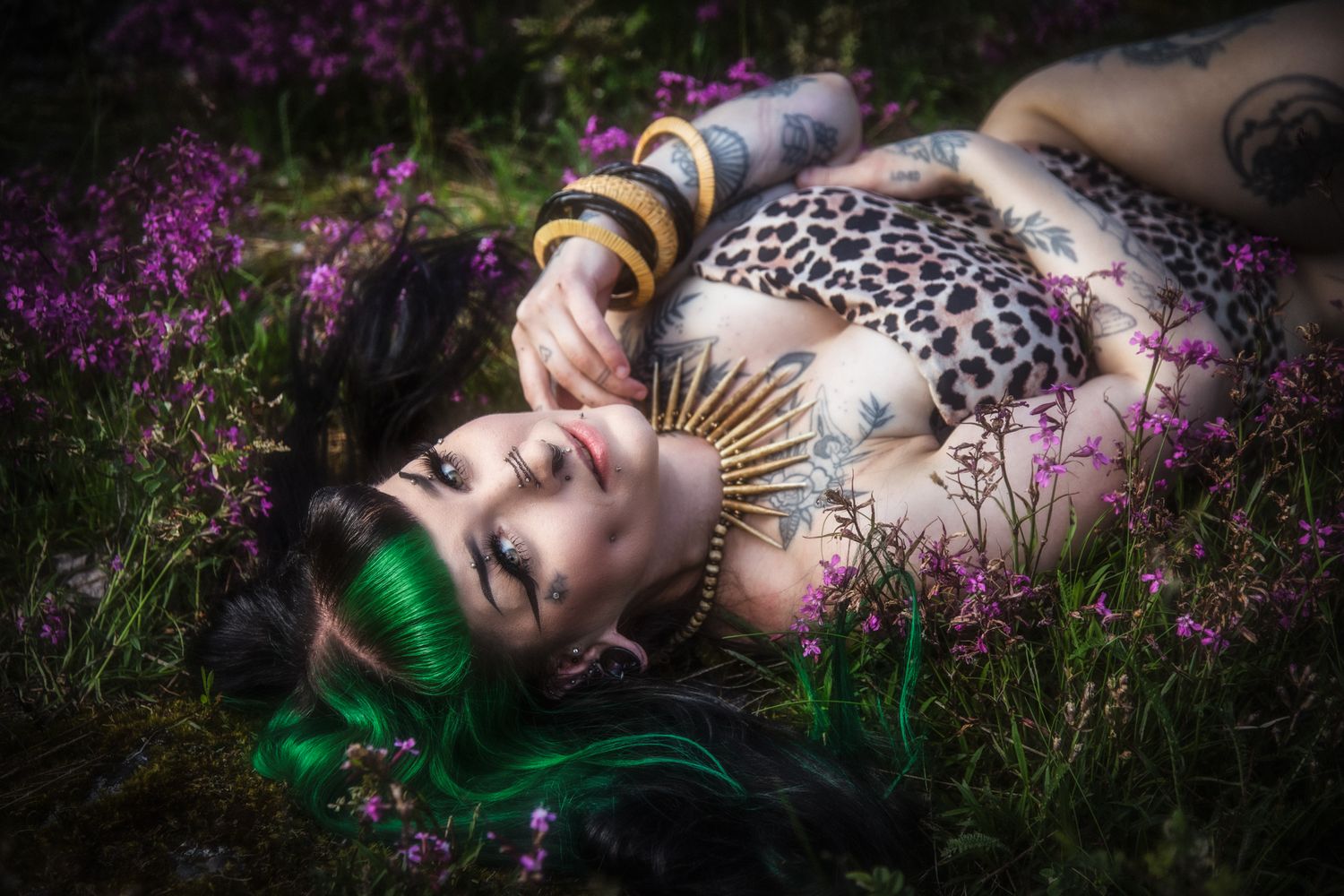 Tattooed woman with green hair lying among purple wildflowers, wearing leopard print top and gold accessories.