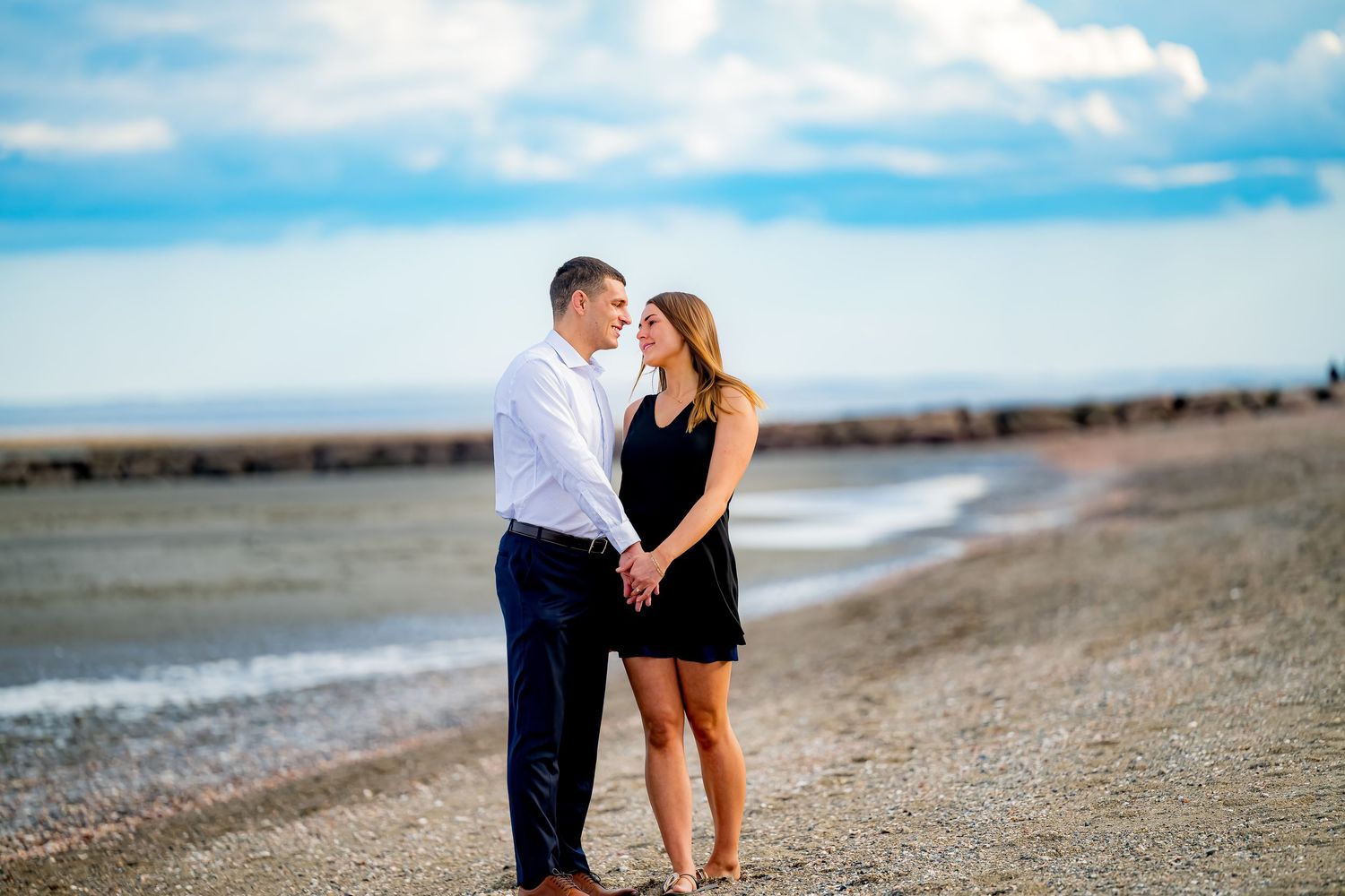 Couple holding hands and gazing at each other on a rocky beach with calm water and cloudy blue sky behind them.