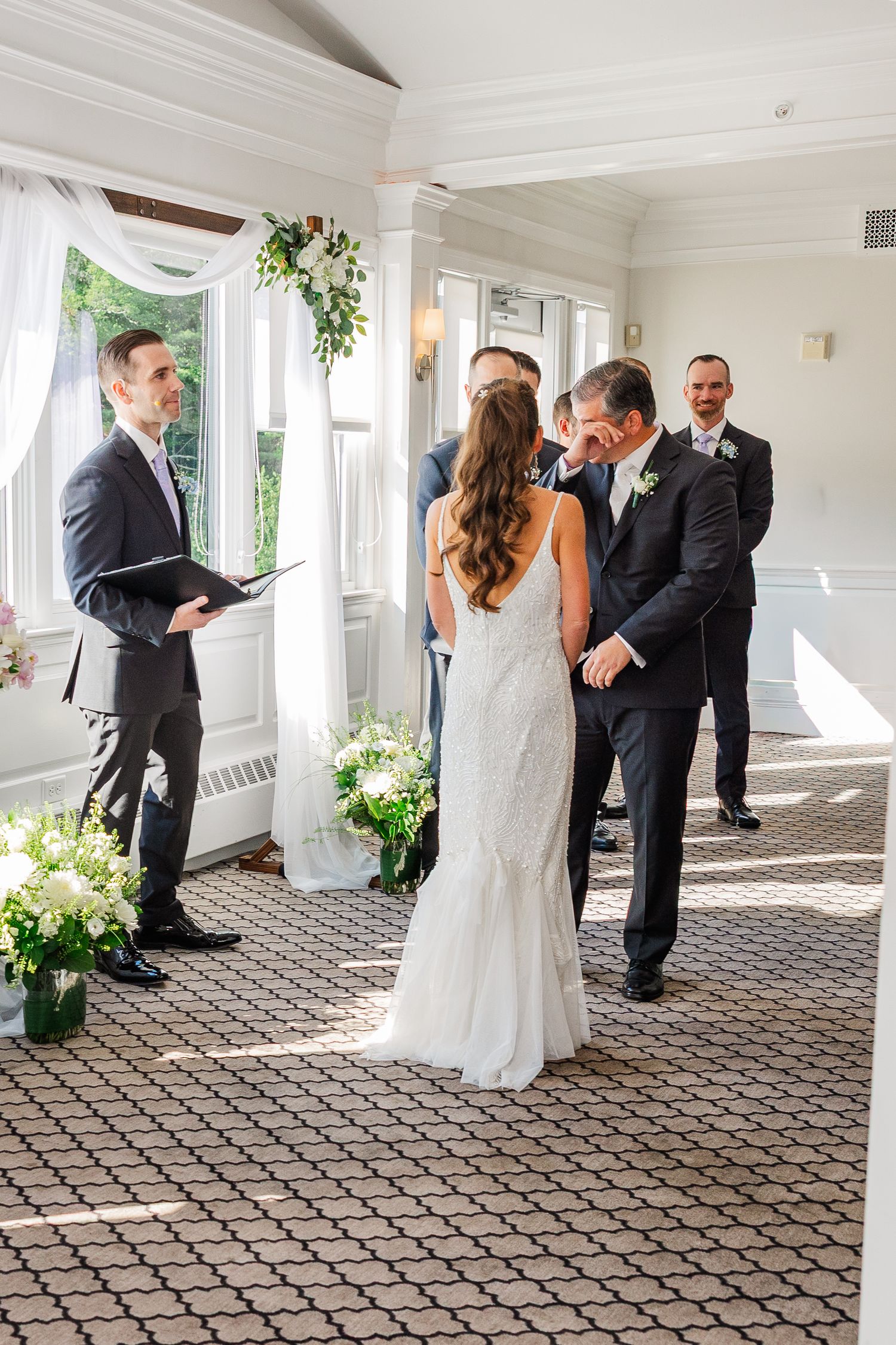 Bride and groom share a kiss during indoor wedding ceremony with officiant and groomsmen in elegant white venue.