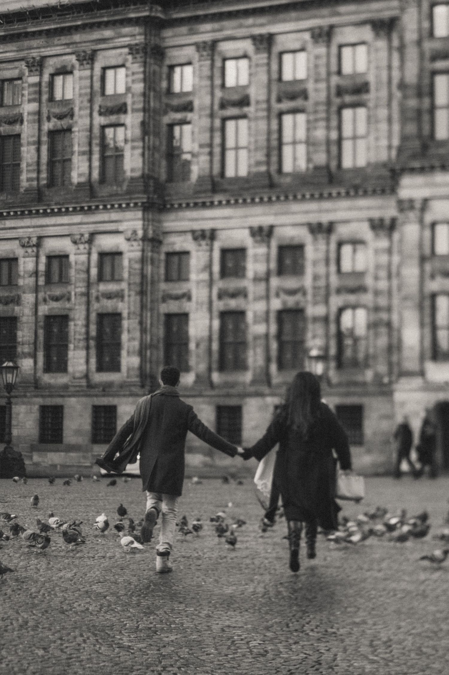 Black and white photo of a couple holding hands and running through a plaza of pigeons in front of Koninklijk Paleis Amsterdam.