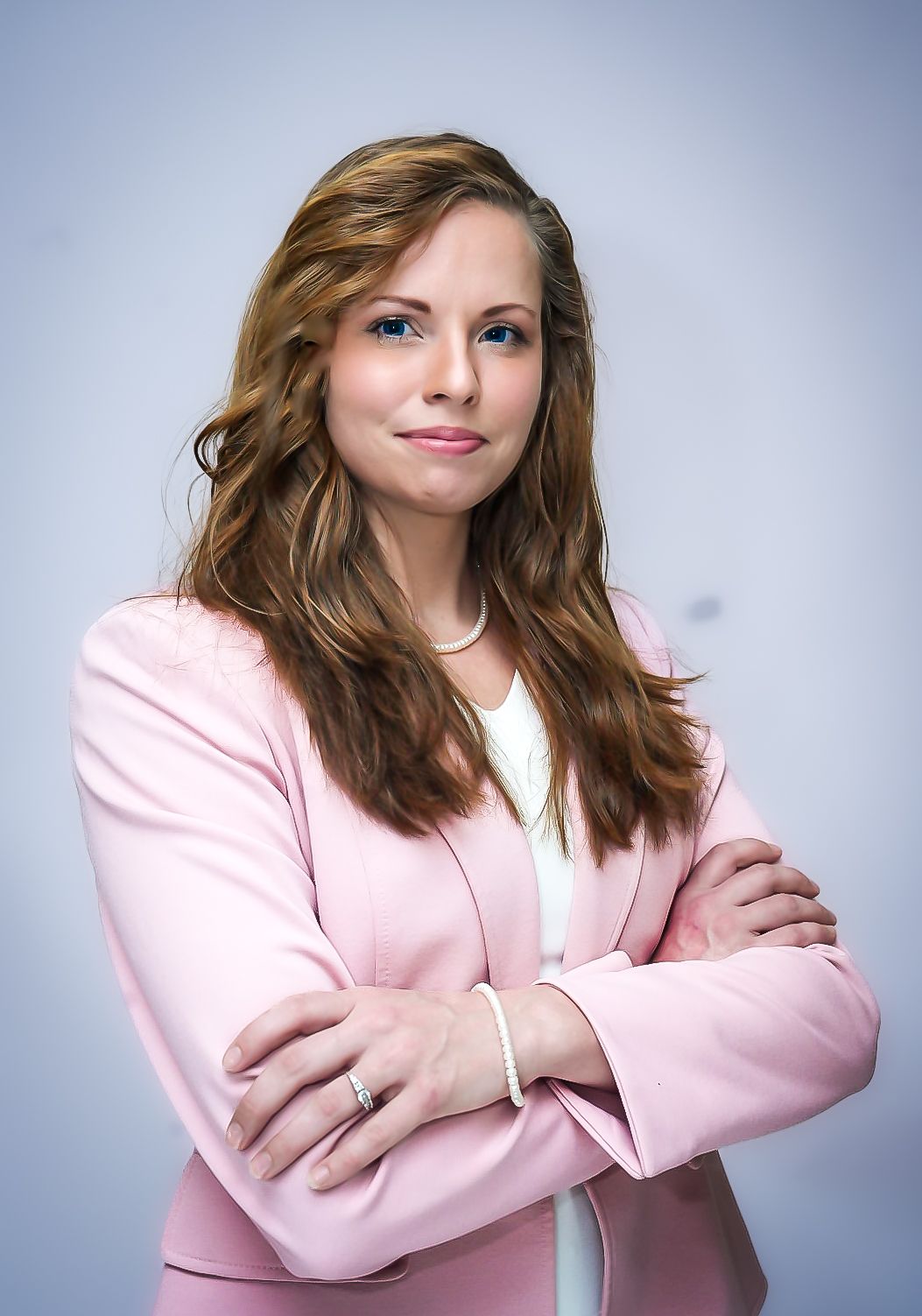 Professional woman in pink blazer with arms crossed, wearing pearl jewelry, posing confidently against light background.