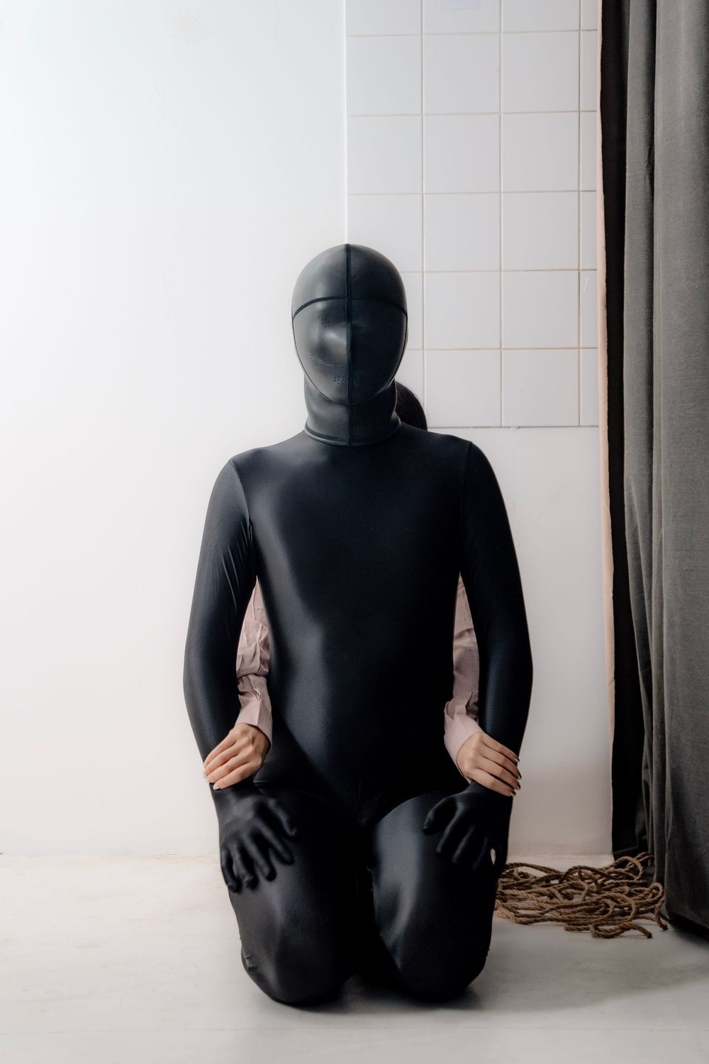 Person wearing a black full-body latex suit and mask kneeling on white floor near a curtain.
