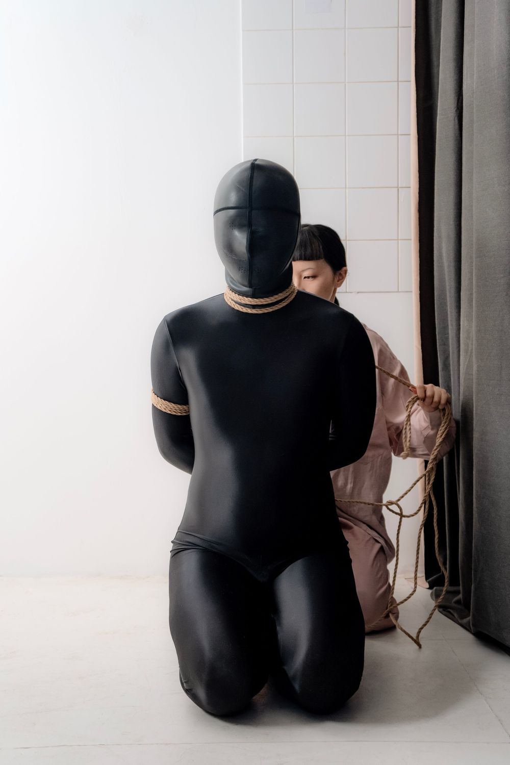 Person in black latex suit kneeling while another ties rope in a white studio setting.