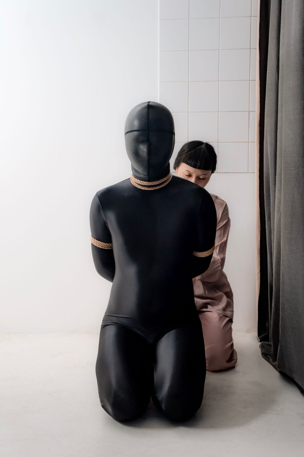Person in black latex suit bound with rope kneeling beside another individual in a beige outfit against white wall.