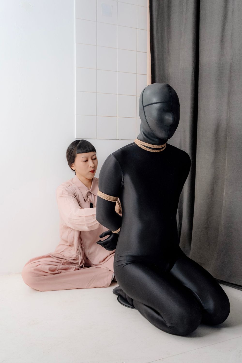 Woman in pink outfit tying rope around person in black full-body suit kneeling on white studio floor.