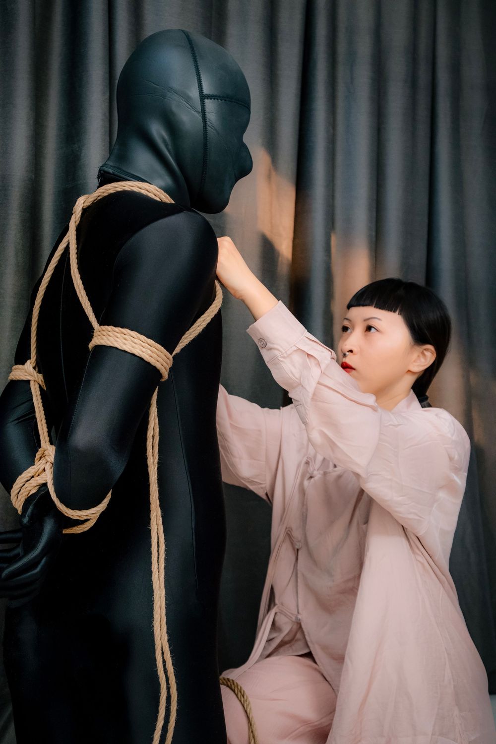 Woman in pink outfit tying rope around a black zentai suit mannequin against gray curtain backdrop.