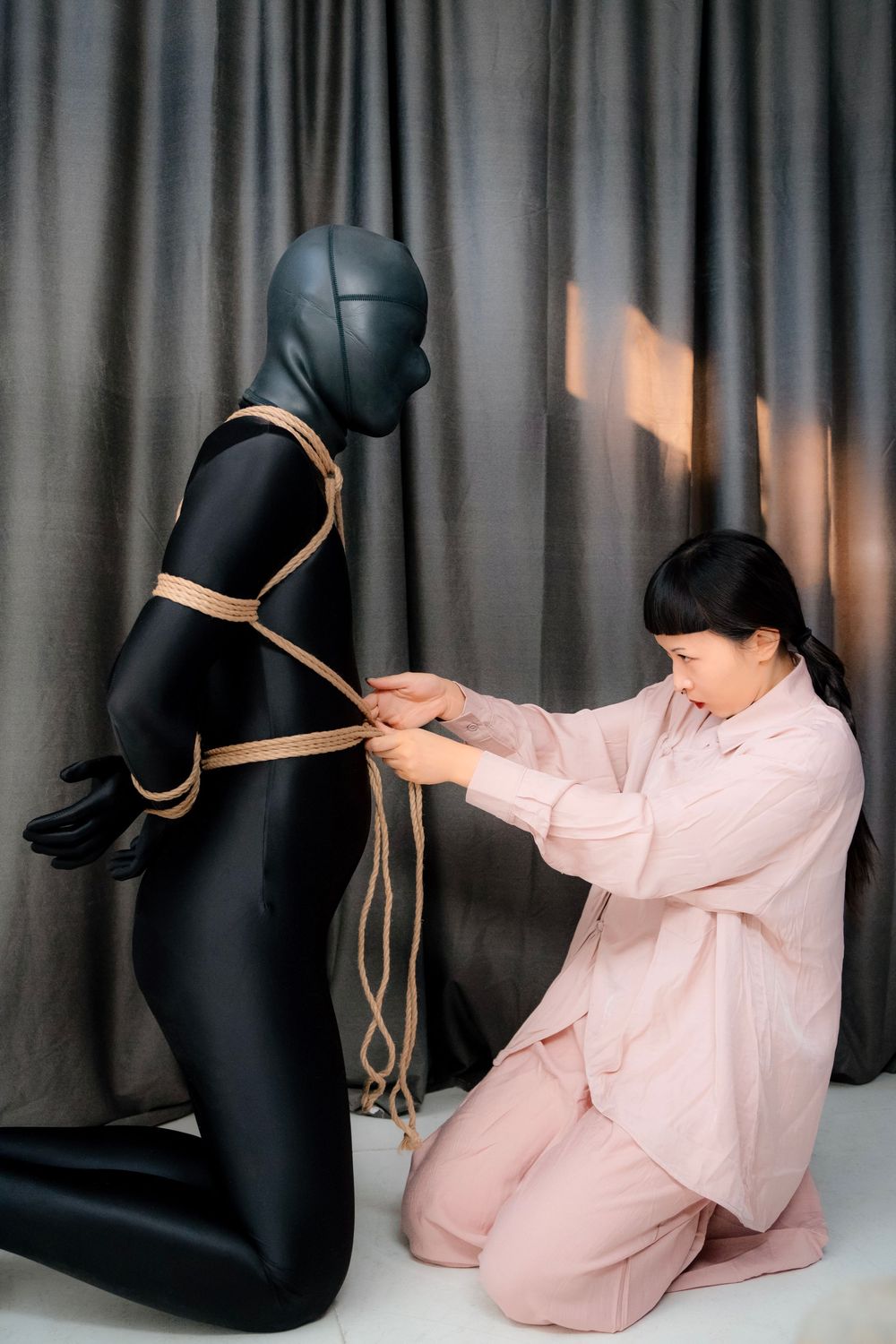 Woman in pink outfit practicing Shibari rope bondage technique on a mannequin in black zentai suit indoors.