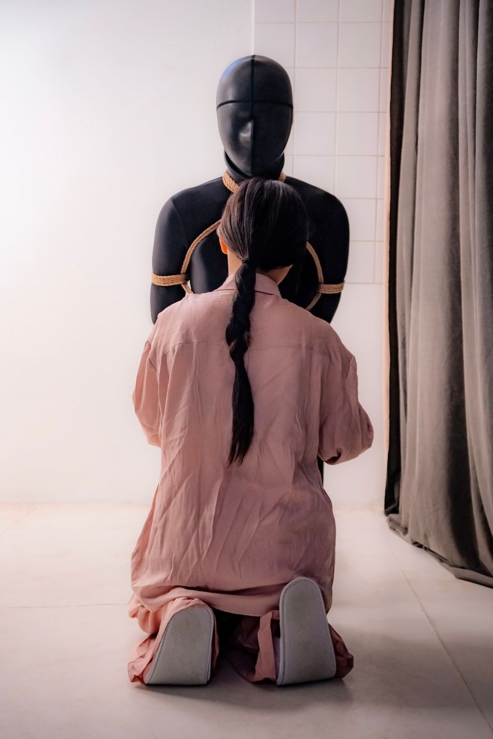 Person kneeling before a black mannequin bound with rope in a white room with curtains.