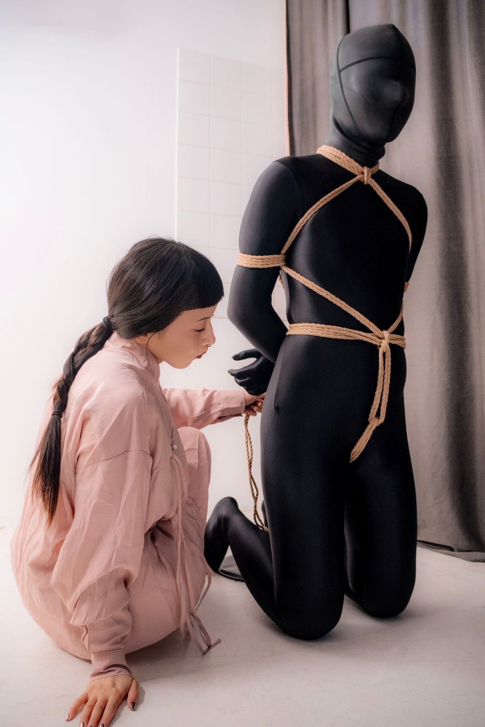 Woman in pink tying rope around a black full-body mannequin in a white studio setting.