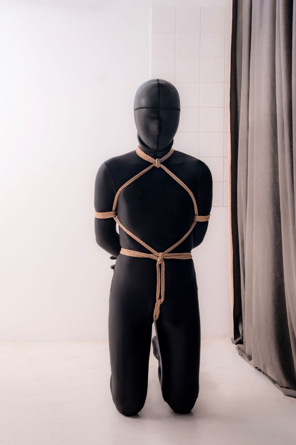 Person in black full-body suit kneeling with decorative rope harness against white tiled wall.