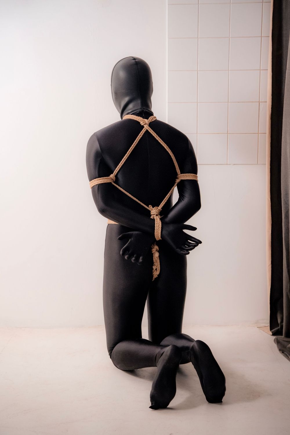 Person in black latex suit kneeling with hands bound behind back with rope against white wall.