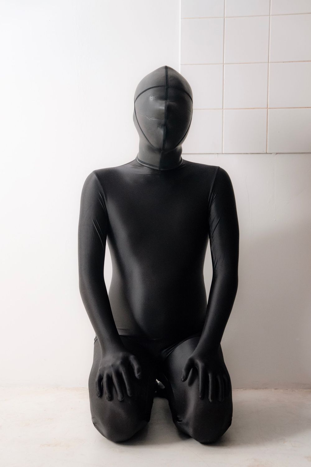 Person wearing a full-body black zentai suit kneeling against a white tiled wall.
