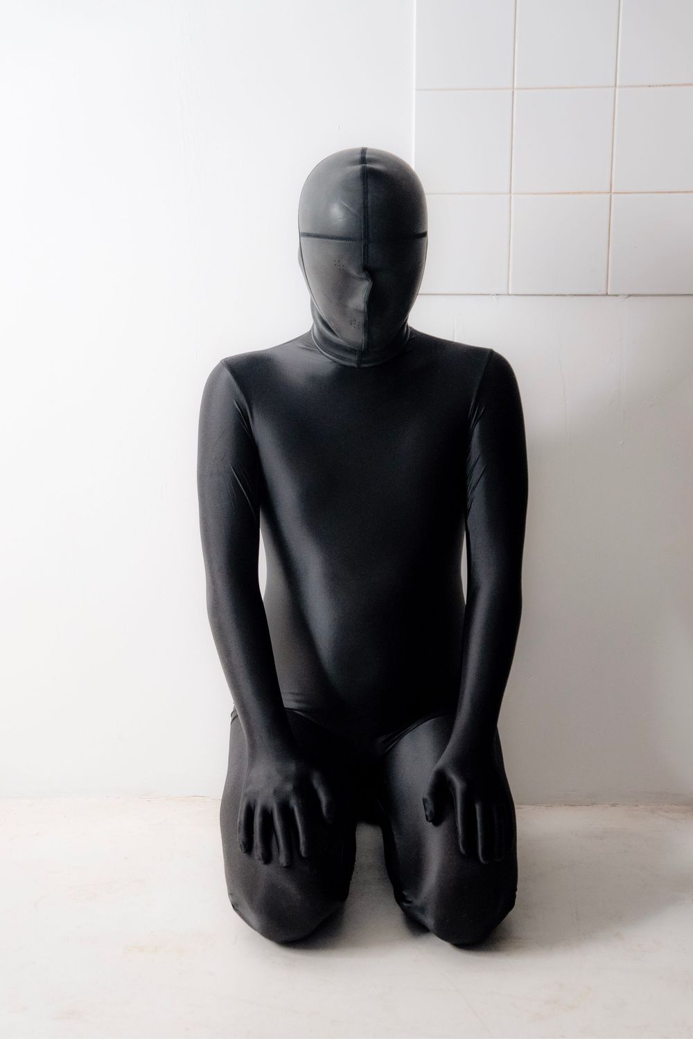 Person in black full-body spandex zentai suit kneeling on white floor against tiled wall.