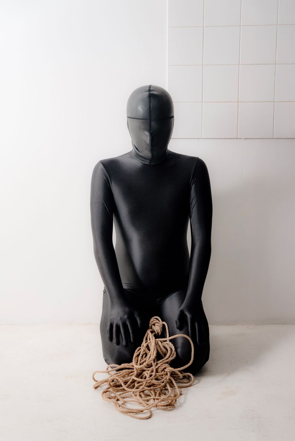 Person in black full-body suit kneeling beside a pile of rope against a white tiled wall.