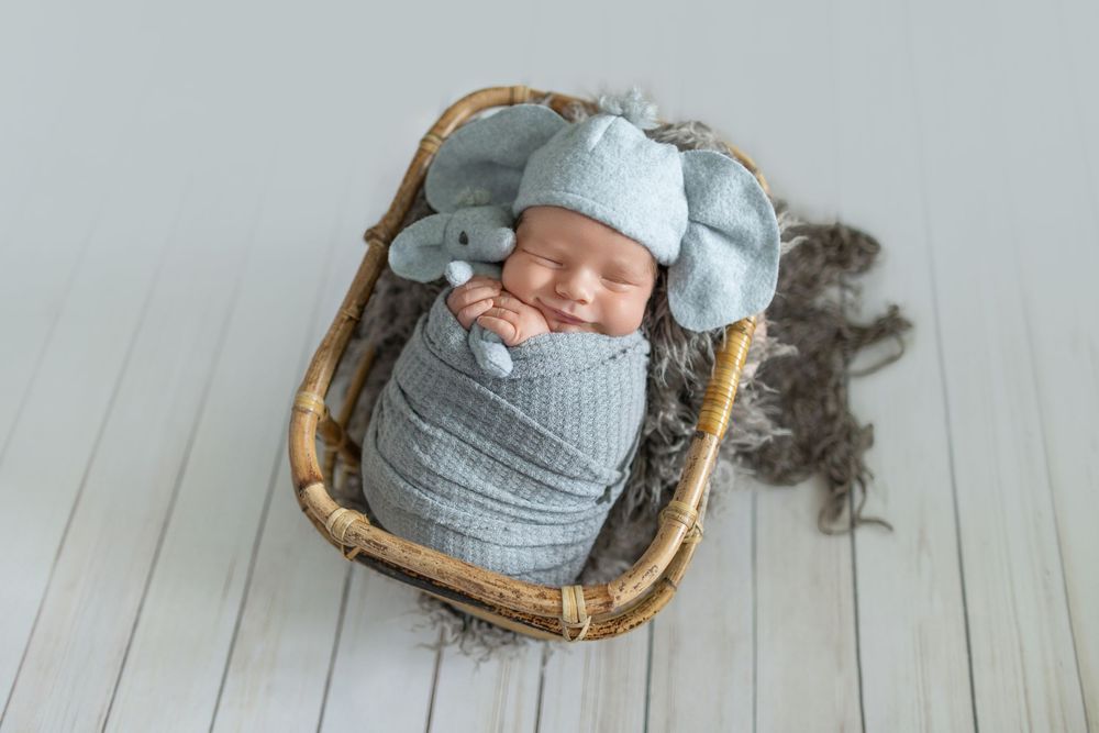 Smiling newborn baby swaddled in blue wrap wearing a knit hat, sleeping peacefully in a wicker basket.