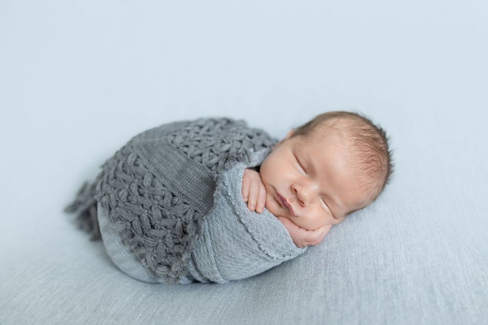 Sleeping newborn baby swaddled in a soft gray knit wrap, posed on a light blue blanket.