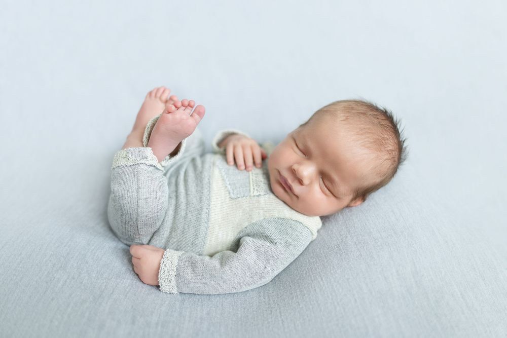 Peaceful newborn baby sleeping in gray romper on soft blue blanket, smiling with tiny feet visible.