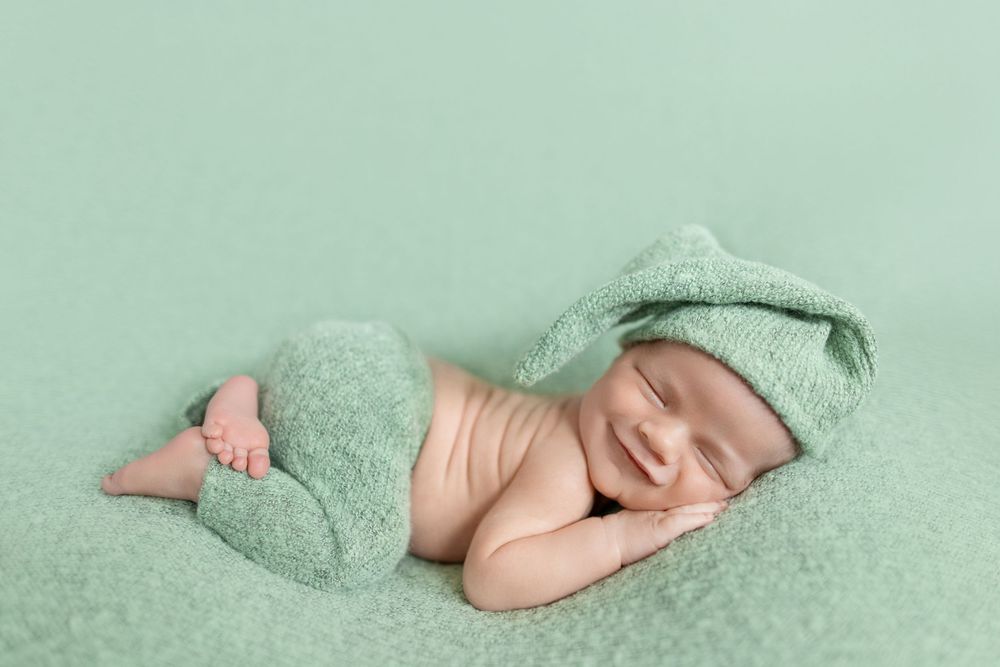 Smiling newborn baby sleeping on mint green blanket wearing a matching knit hat and pants.